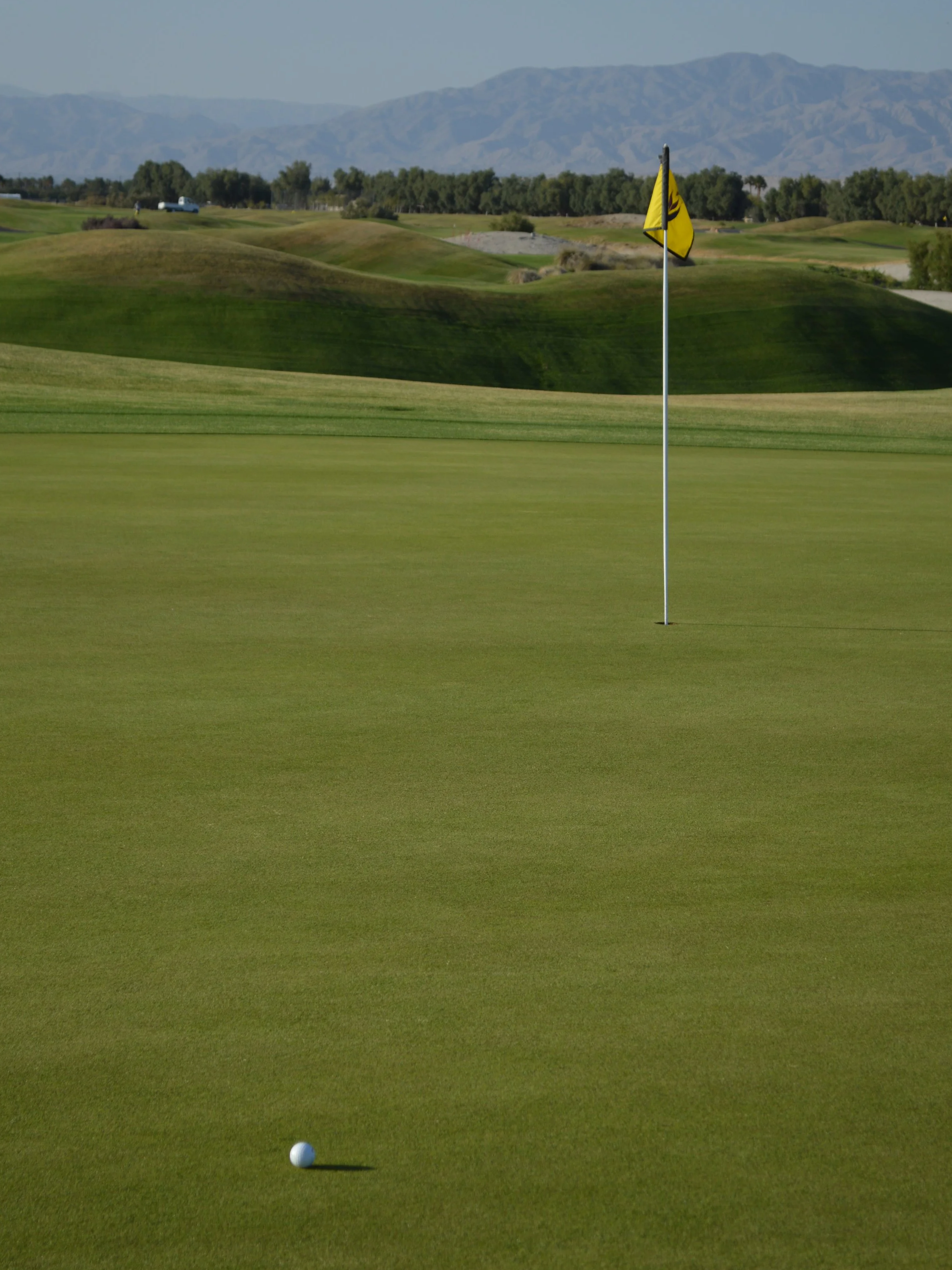 A golf ball on the putting green near the hole with a yellow flag in Palm Springs CA, with a golf course and mountains in the background.