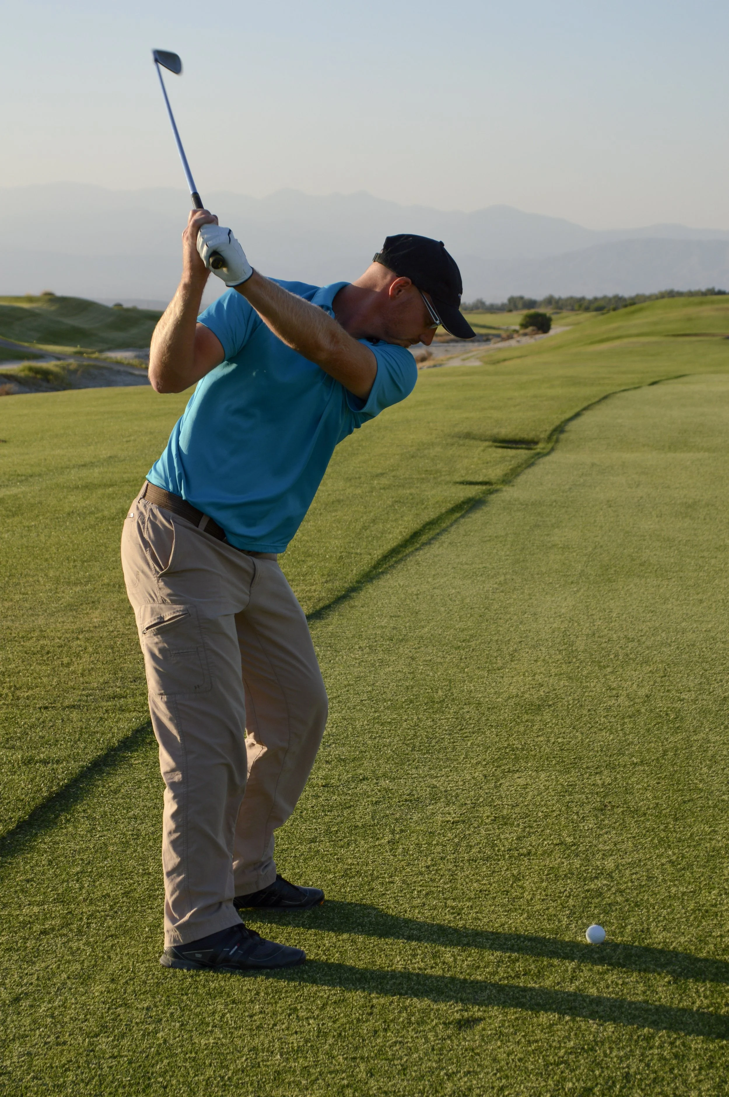 PGA Master Professional Dave Cahill student is mid-swing on a golf course, preparing to hit a golf ball with a club. The course is lush and green with mountains visible in the background, under a clear sky.