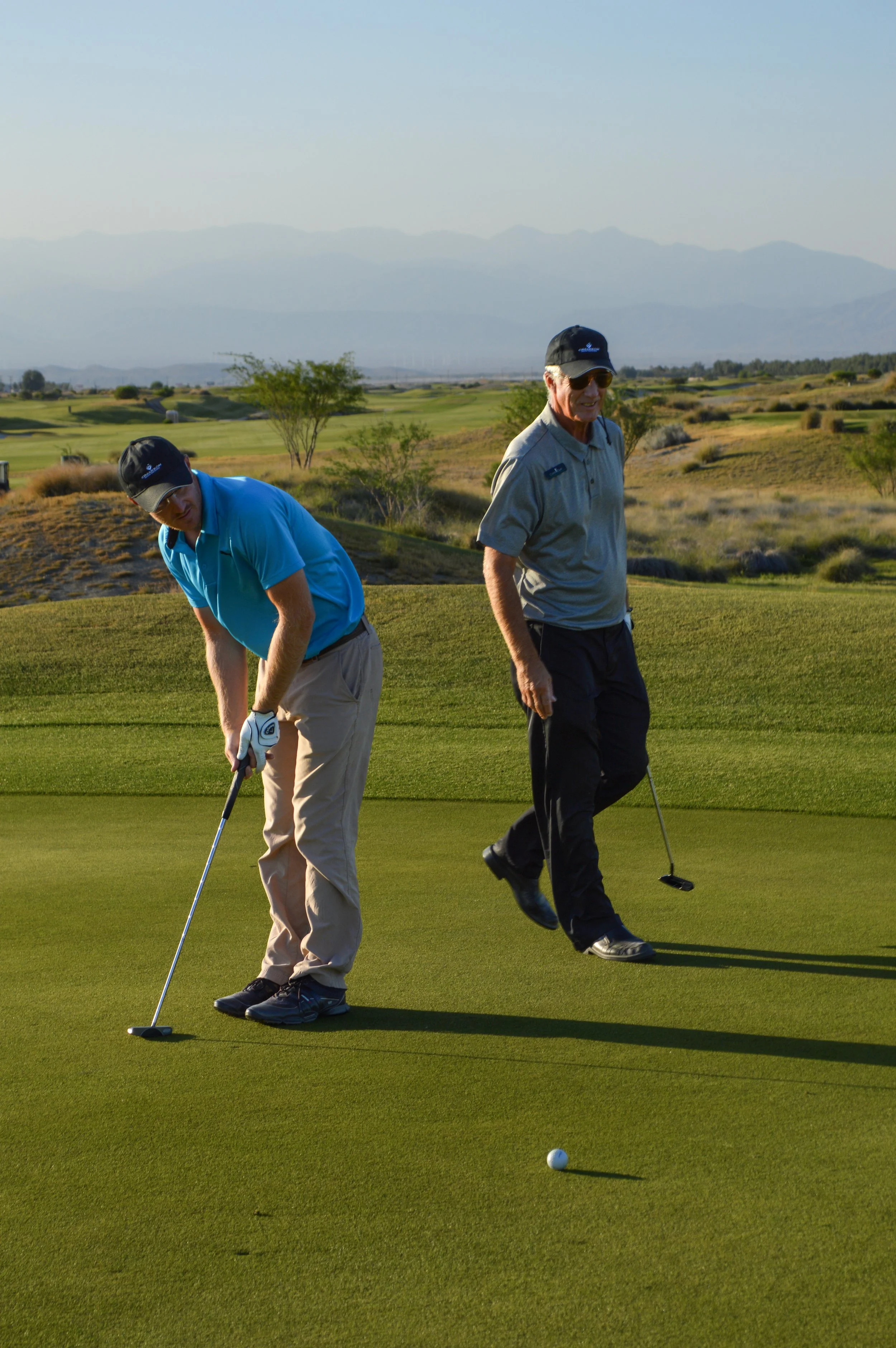 Palm Springs CA men playing golf on a sunny course, one preparing to putt the ball while the other walks nearby with a golf club, green fairways, trees, and mountains in the background.