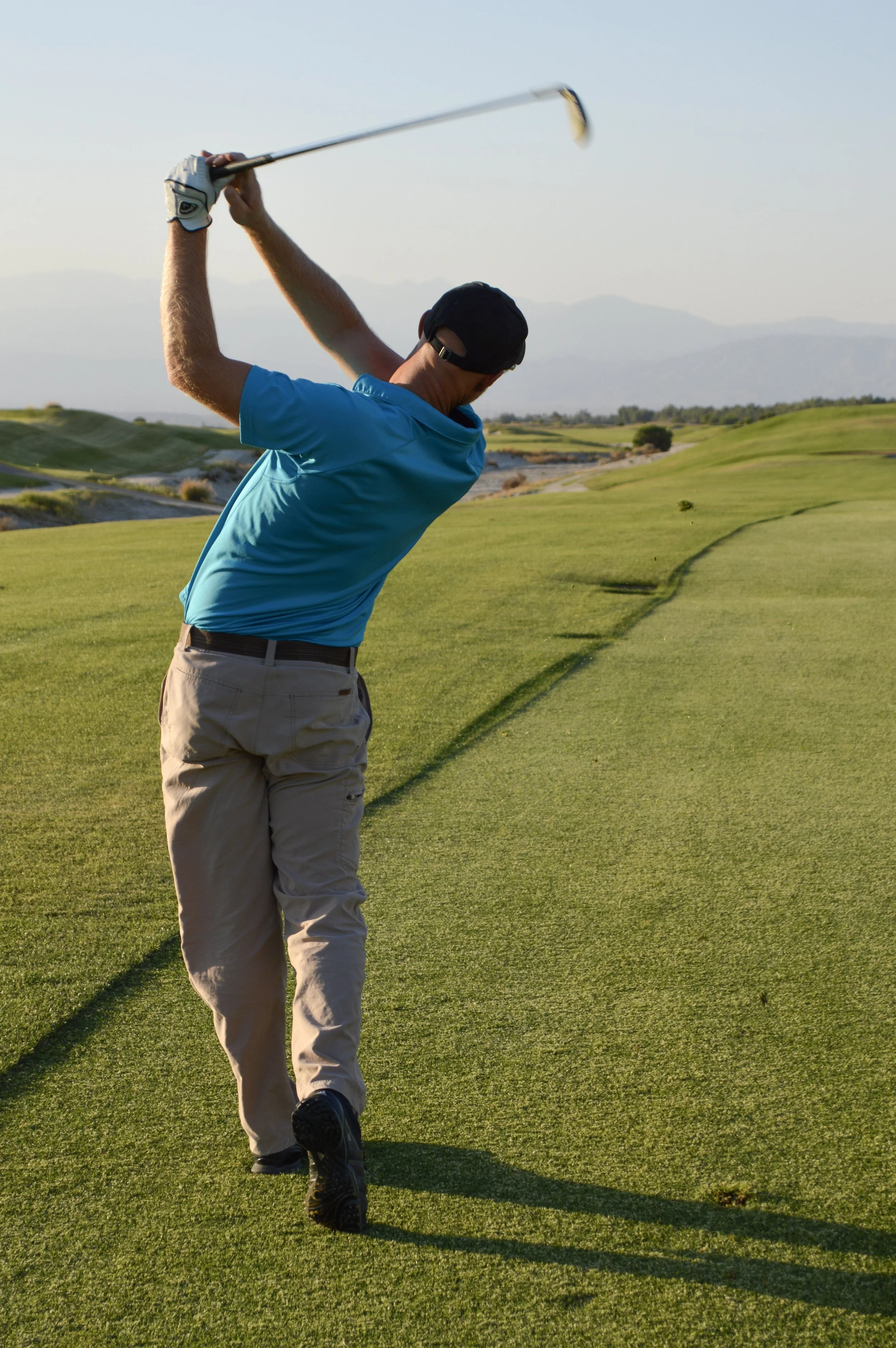 Palm Springs golf student in a blue shirt and beige pants is playing golf on a lush green course, swinging a golf club with mountains in the background, during the daytime.
