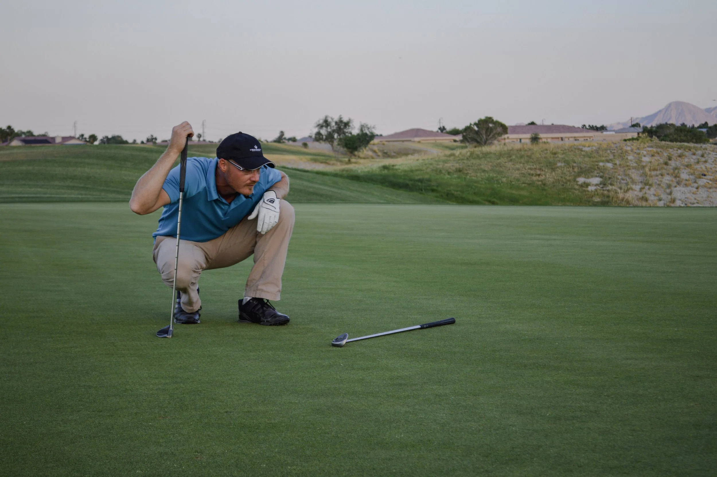 Palm Springs golf student in a blue polo shirt, khaki pants, and a black cap practicing putting on a golf green, crouching down with his hand on a golf club, with a golf ball on the ground in front of him.