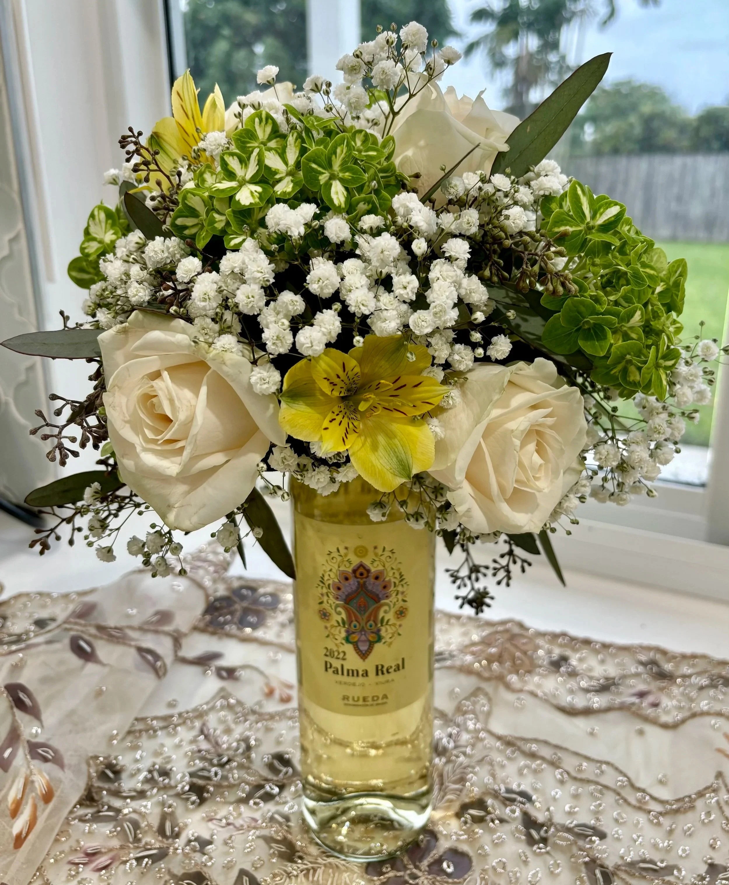 White roses, baby's breath and yellow alstroemeria arranged around a wine bottle