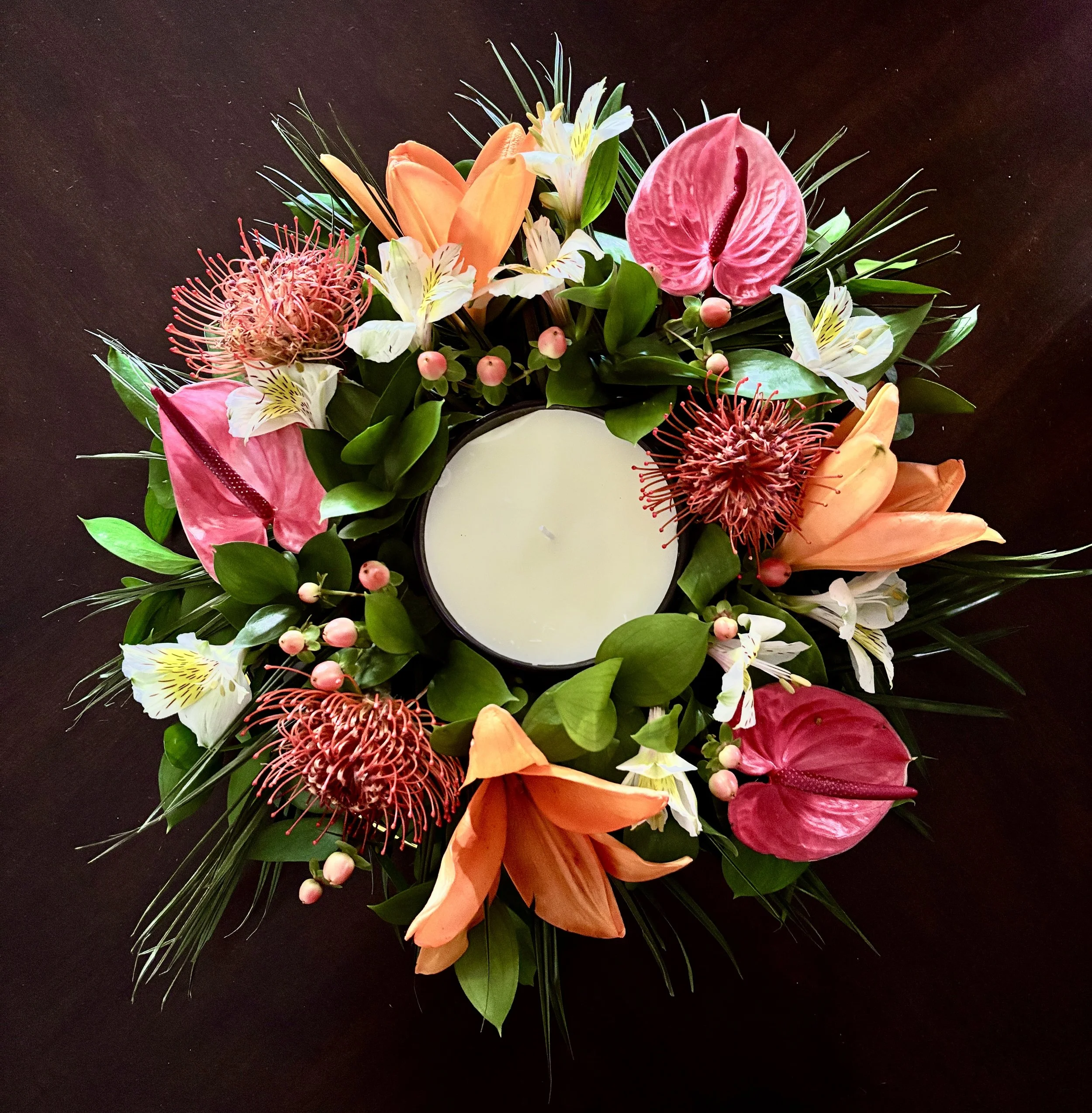 Round centerpiece of orange lilies, pink anthuriums and pincushion protea around a white pillar candle