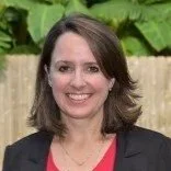 A smiling woman with shoulder-length brown hair, wearing a black blazer and a red top, standing outdoors in front of a wooden fence and green foliage.