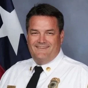 Portrait of a man in a white police uniform with a gold badge, smiling, with a partial view of the American flag in the background.