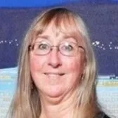 A woman with long blonde hair and glasses, smiling in front of a blue background with some string lights.
