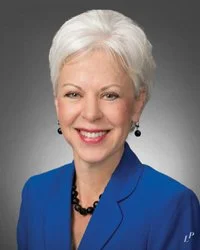 Portrait of an elderly woman with short white hair, wearing a blue blazer and black necklace, smiling against a gray background.