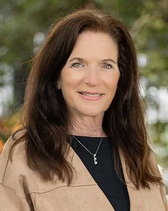 Woman with long brown hair smiling outdoors, wearing a black top and tan jacket, with blurred green trees in the background.