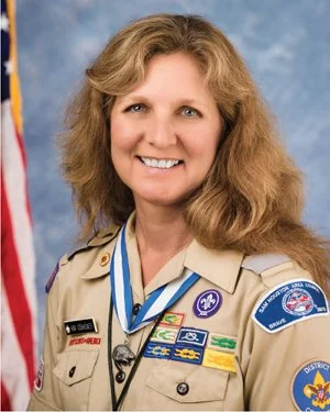 Portrait of a smiling woman in a Girl Scouts uniform with patches and medals, standing in front of an American flag.