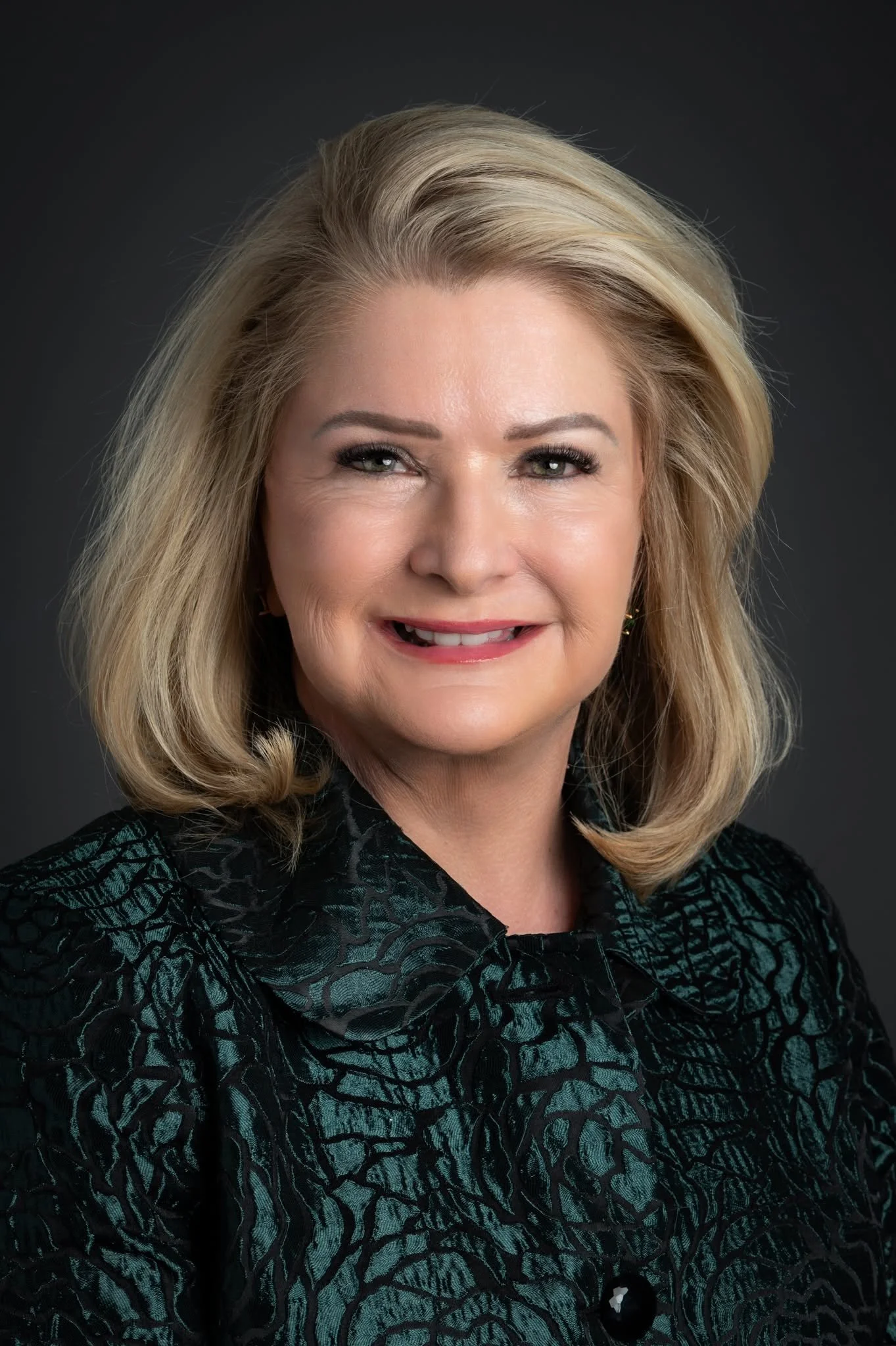 Close-up portrait of a smiling middle-aged woman with blonde hair, wearing a dark patterned top, set against a dark background.
