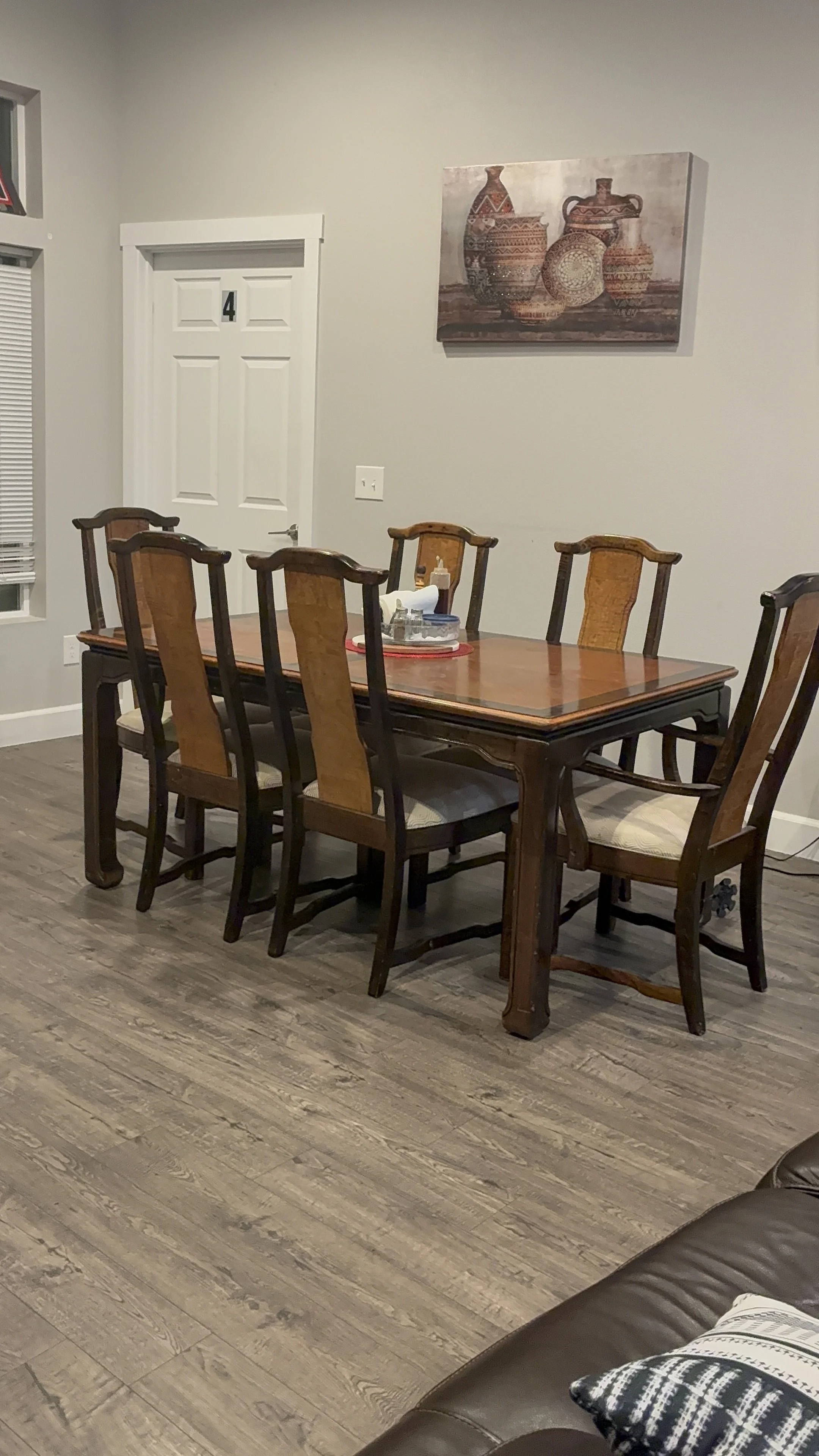 A dining room with a wooden table and six matching wooden chairs with beige cushioned seats. Above the table is a painting of traditional pottery. To the right, part of a dark brown leather couch with a patterned pillow is visible.