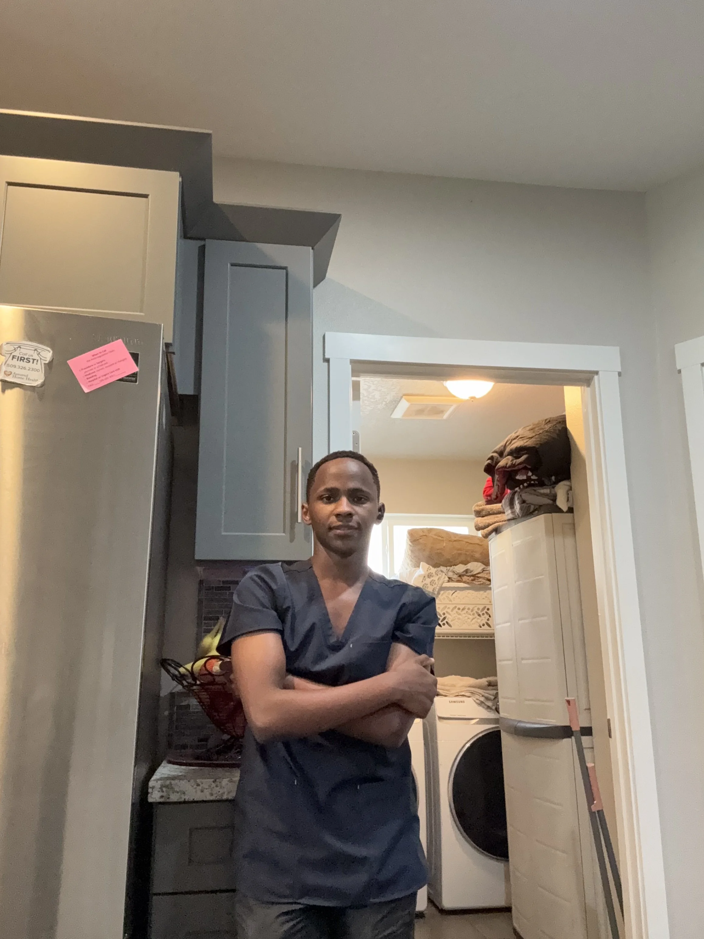 A man with short hair and wearing dark scrubs stands with arms crossed in a laundry room, with cabinets, a washing machine, and a closet with folded clothes in the background.