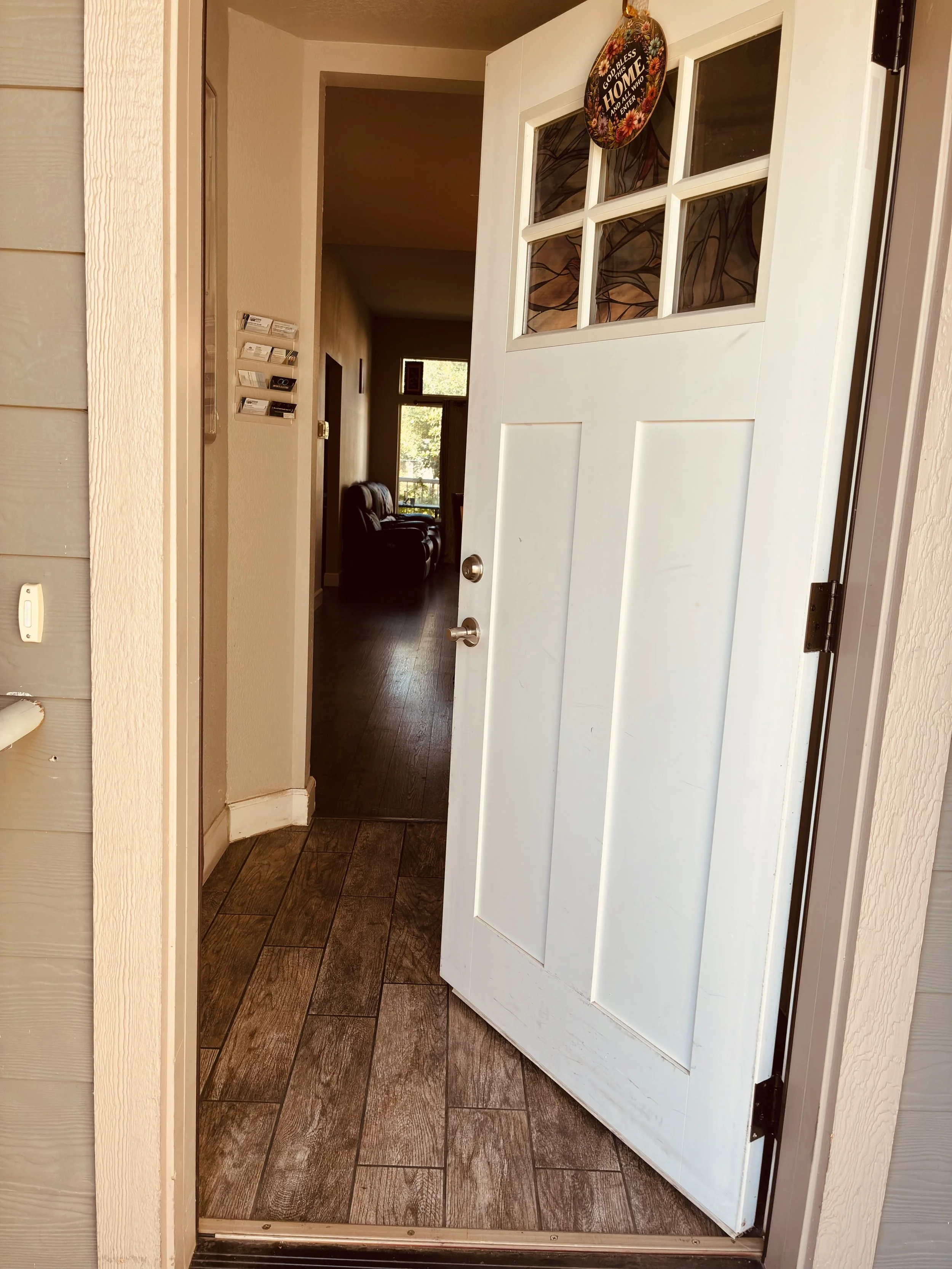 View of a front door opening into a hallway with wooden flooring and a sitting area in the background. A decorative wall piece hangs on the door with a floral design and the words 'God bless this home and everyone who enters it'.