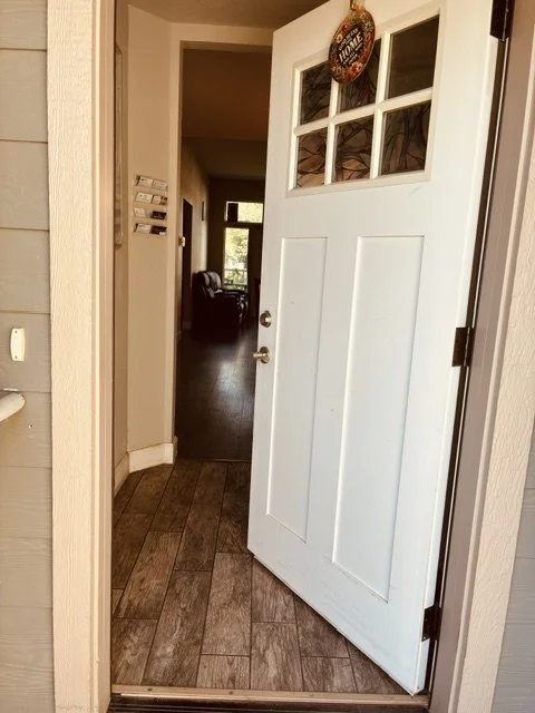 The front door is open, revealing a hallway with a dark wood floor leading to a room with a window and a chair inside. The door has a holiday wreath hanging at the top.