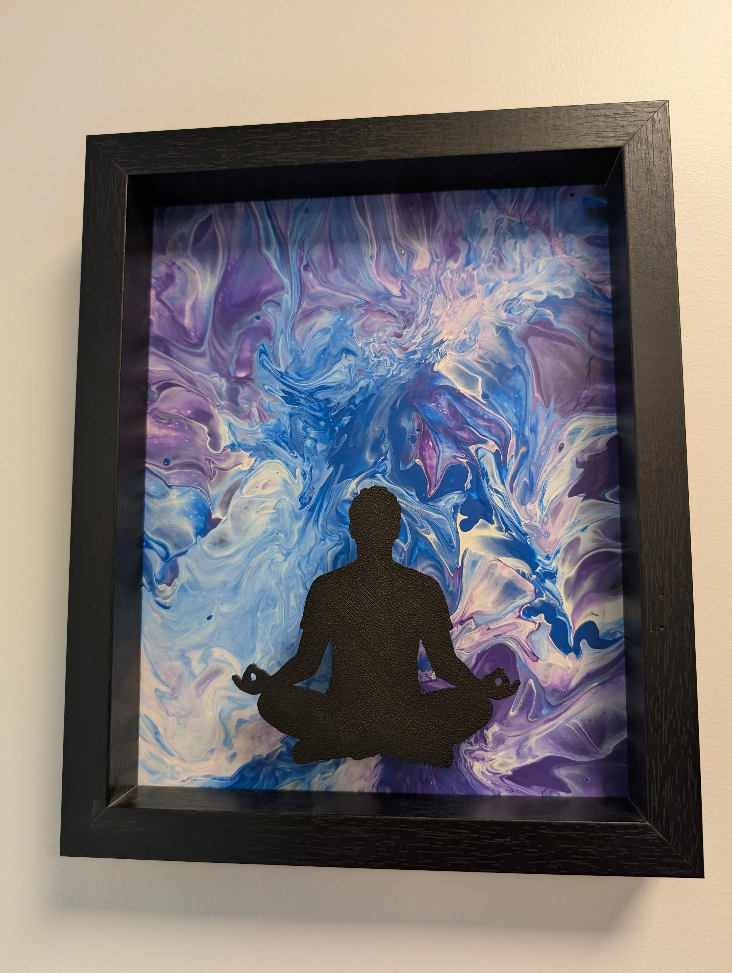 A black framed paintings blues, purples, whites, and greys with a black silhouette in the foreground meditating.