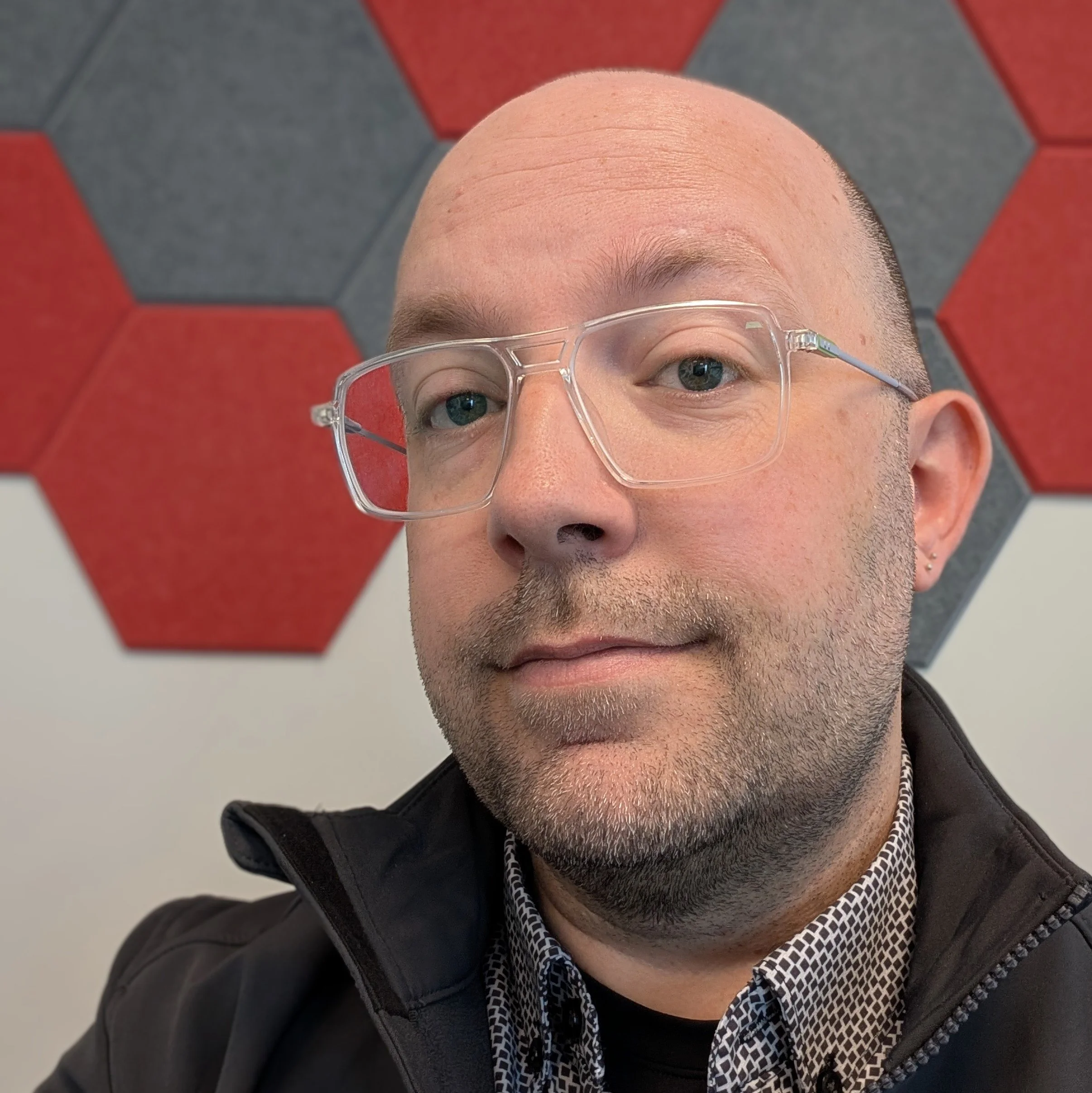 Close-up of a man's face focusing on his forehead, eyes, glasses, and lower face. He has a shaved head, facial stubble, and is wearing a black jacket with a patterned shirt and a chain necklace. The background features a wall with hexagonal tiles in gray and red colors.