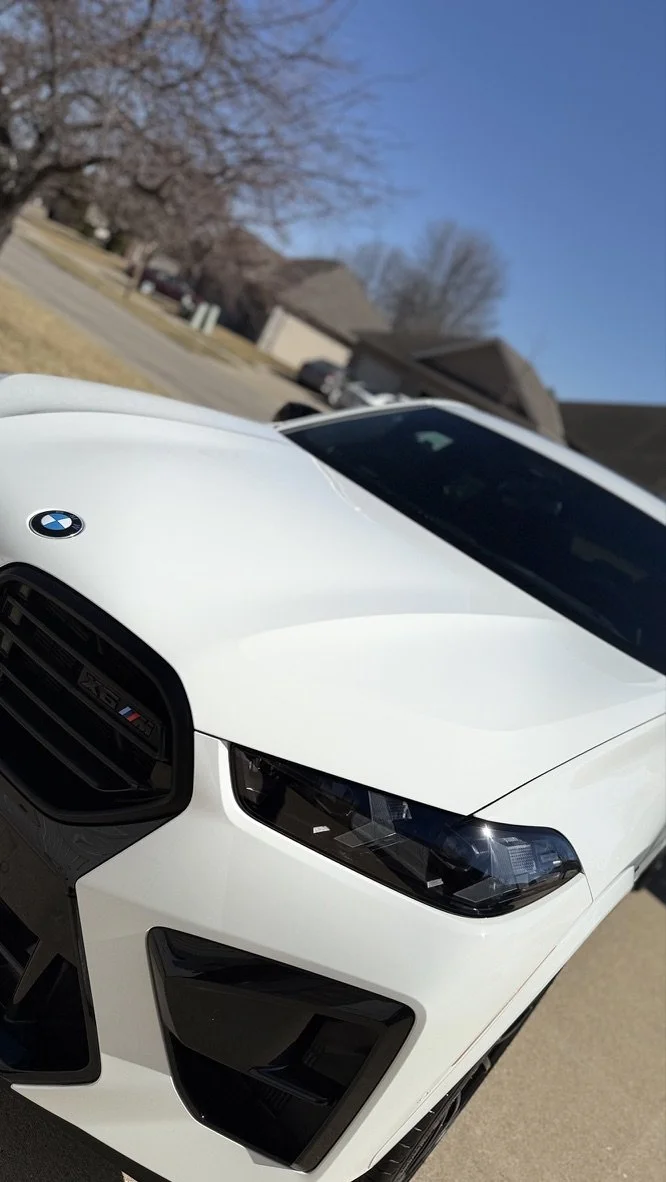 Close-up of a white BMW car parked outside, with trees and houses in the background under a clear blue sky.