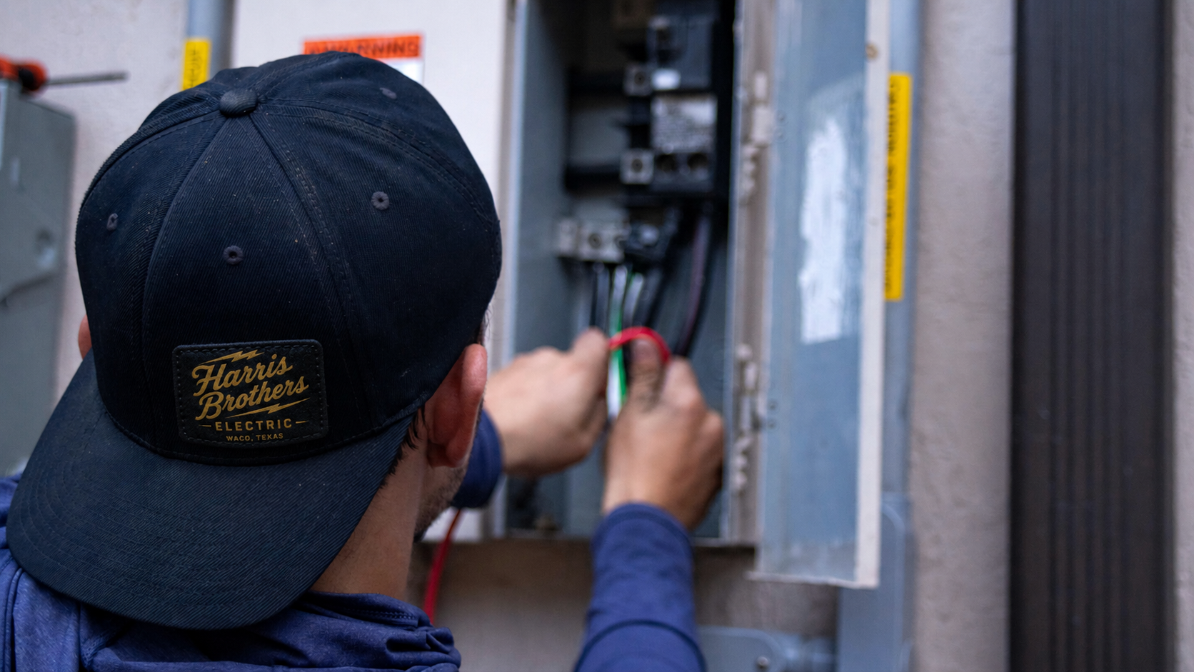 A person wearing a black Harris Brothers Electric cap working on an electrical panel with wires.