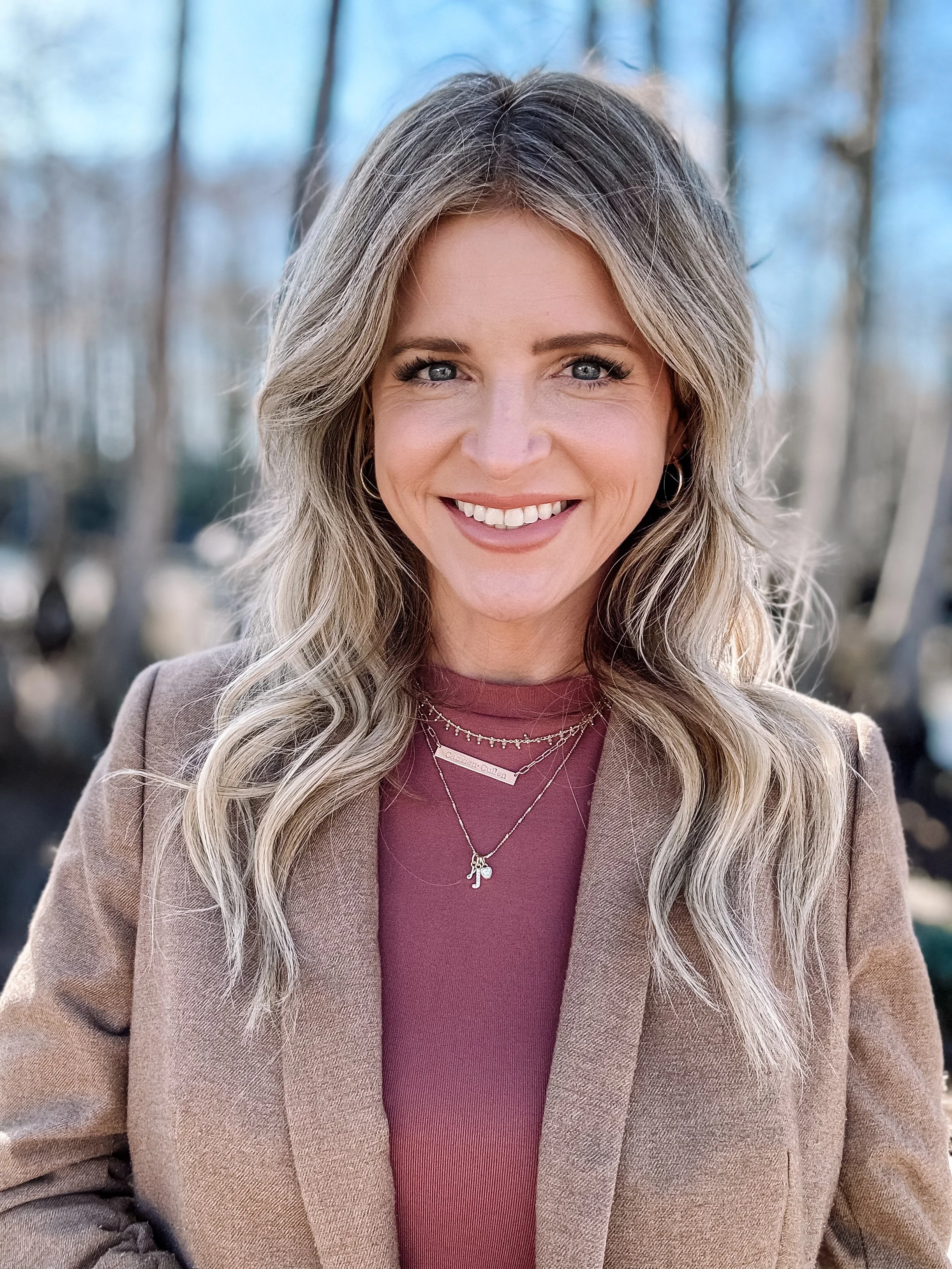 A woman with long, wavy blonde hair and blue eyes smiling outdoors. She is wearing a beige coat and a maroon top with layered necklaces. The background shows a blurred, leafless outdoor setting.