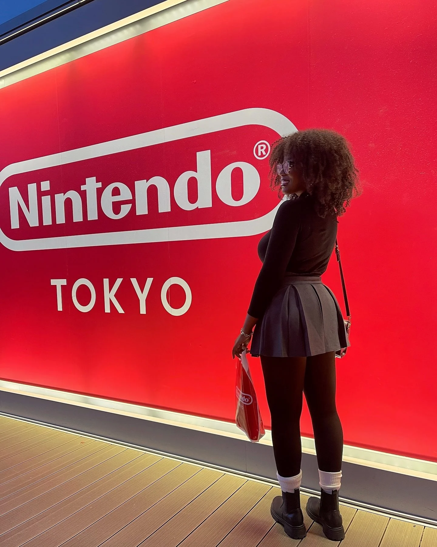 A woman with curly hair, wearing glasses, a black long-sleeve top, a pleated skirt, black tights, and boots, standing in front of a large red wall with a Nintendo and Tokyo logo.