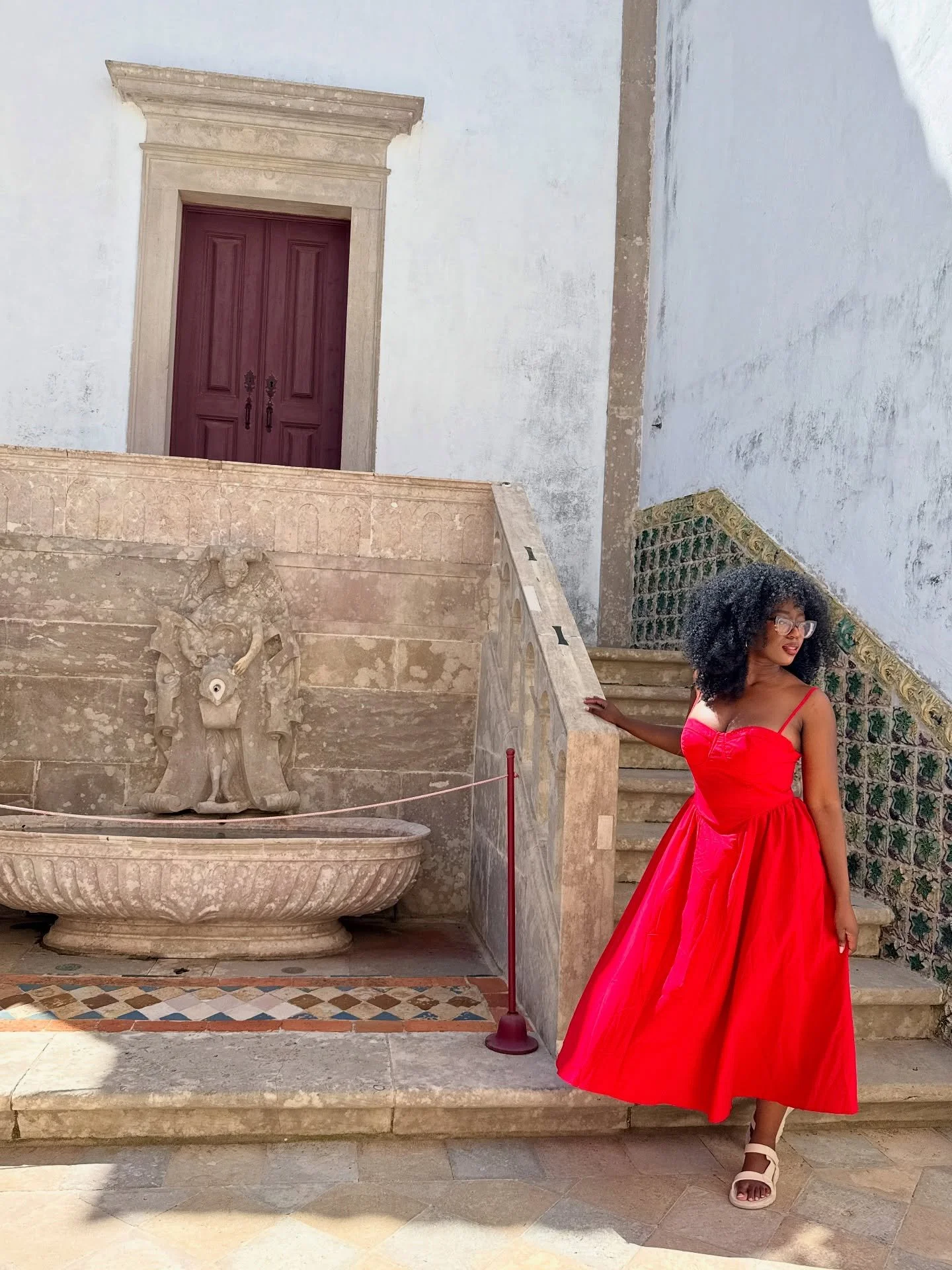 A woman in a bright red strapless dress with a full skirt stands next to a stone fountain and staircase at a historical site. She is wearing glasses, has curly black hair, and is posing with her left hand on the staircase railing.