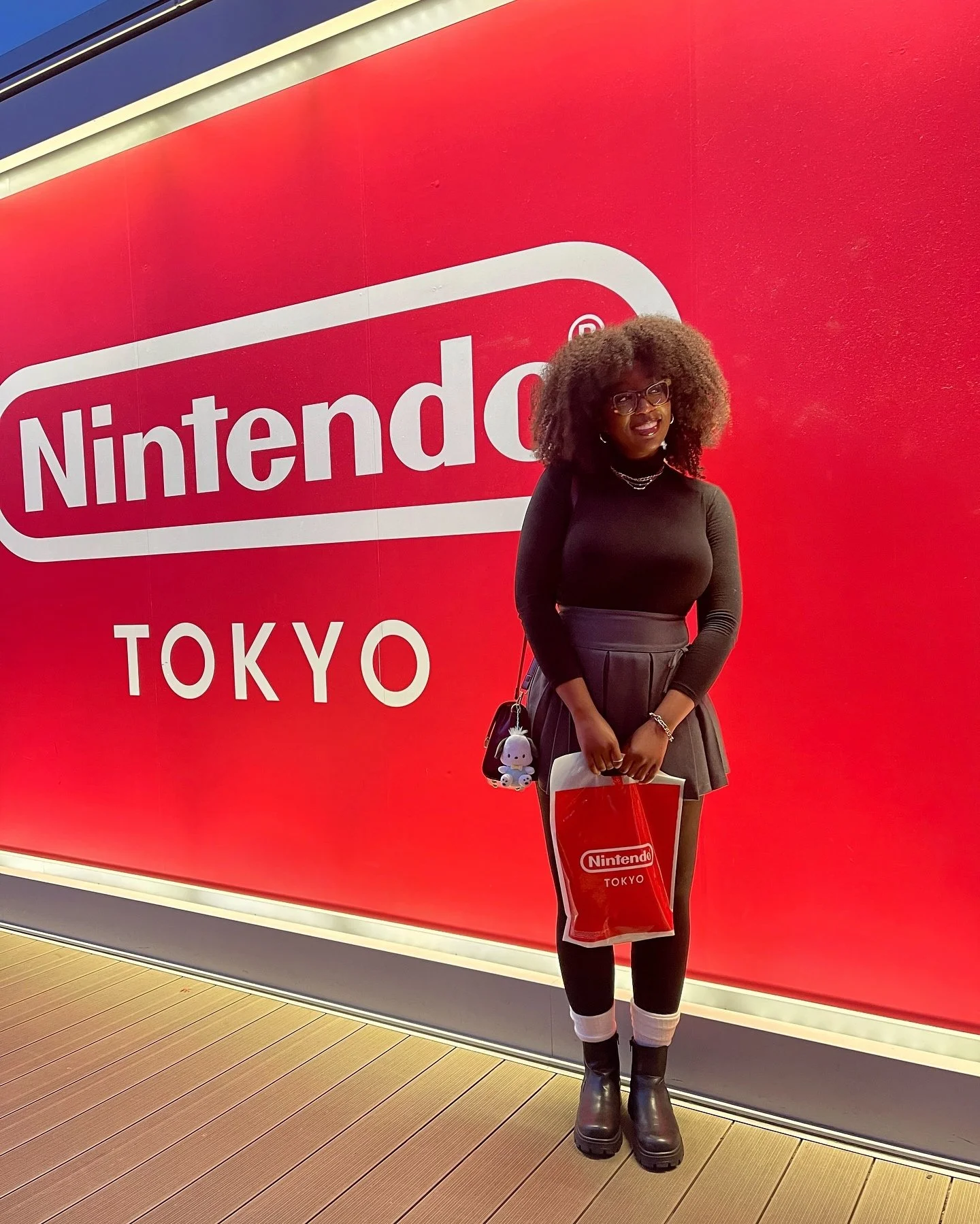 A woman standing in front of a red wall with the Nintendo logo and the word Tokyo. She is holding a red shopping bag with the Nintendo logo, wearing black boots, a black long-sleeve top, a gray skirt, and glasses. She has curly hair and is smiling.