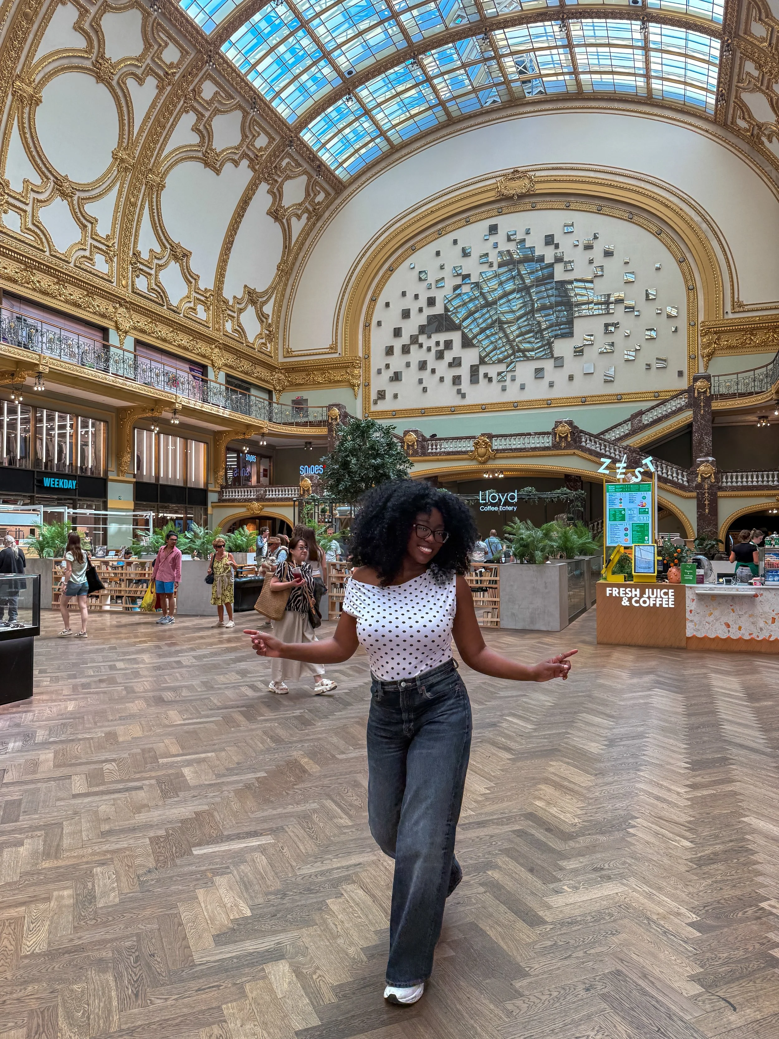 Woman with curly hair, glasses, and a polka dot sleeveless top dancing inside a large, ornate shopping mall with a glass ceiling, gold detailing, and multiple stores and shoppers in the background.