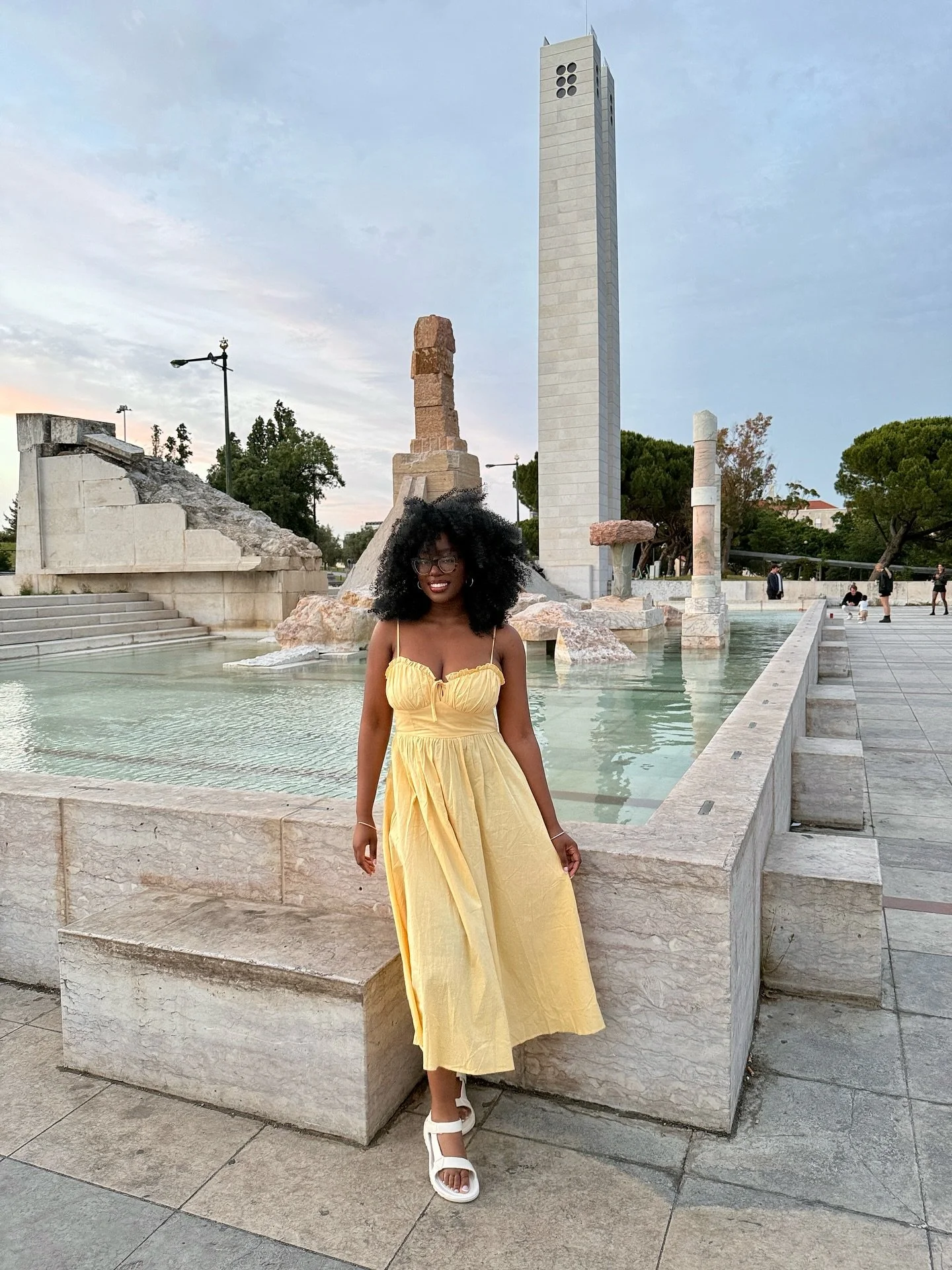 A smiling woman in a yellow dress and white sandals standing in front of a historic fountain and monument in an outdoor public space at dusk.
