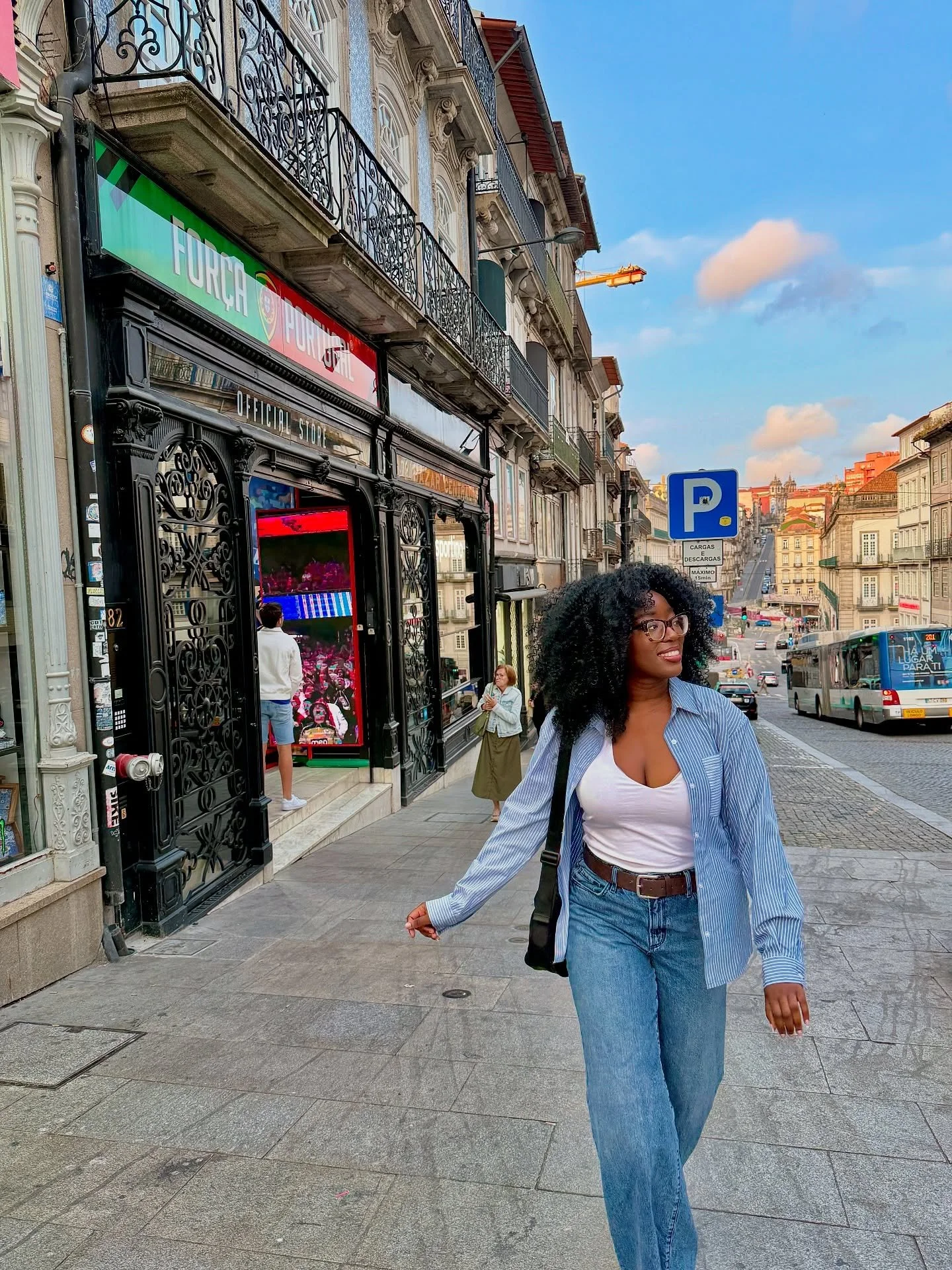 A woman with curly black hair, glasses, a white top, a blue striped shirt, and jeans walking on a city sidewalk.