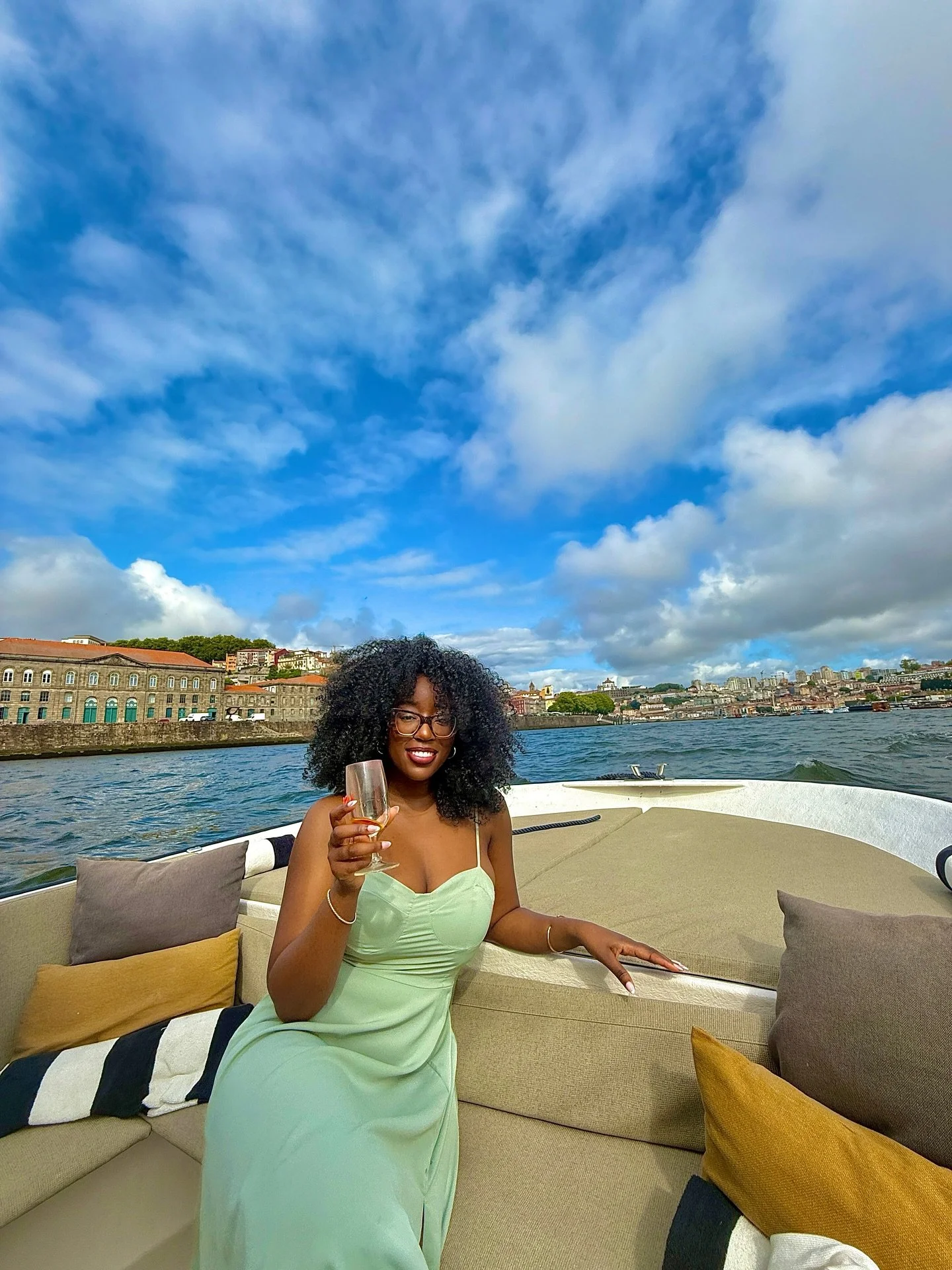 A woman with curly hair and glasses sitting on a boat, holding a glass of champagne, with city buildings and a cloudy sky in the background.