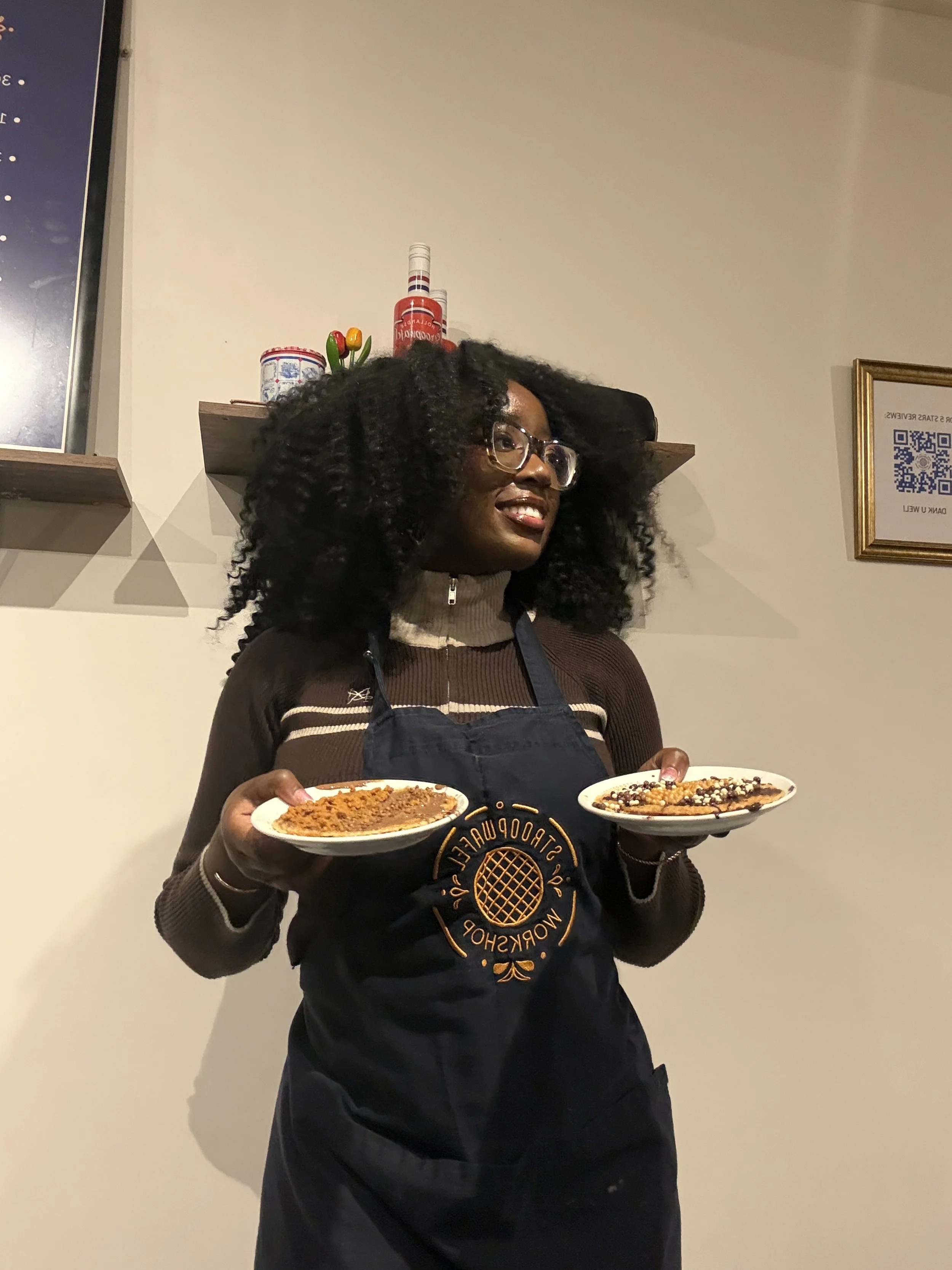A woman with curly black hair, glasses, and a brown sweater, wearing a black apron, holding plates of dessert while smiling in a restaurant.