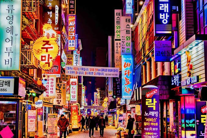 Night view of a busy street in South Korea with numerous illuminated neon signs in Korean. People are walking along the street, and various shops and businesses line the street.