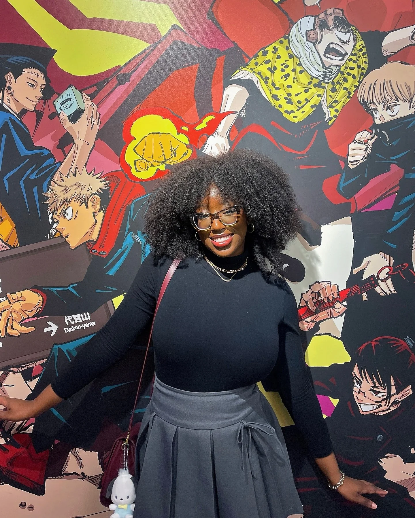 A woman with curly black hair, glasses, and wearing a black top with a gray skirt, standing in front of a colorful anime-style mural featuring multiple characters.