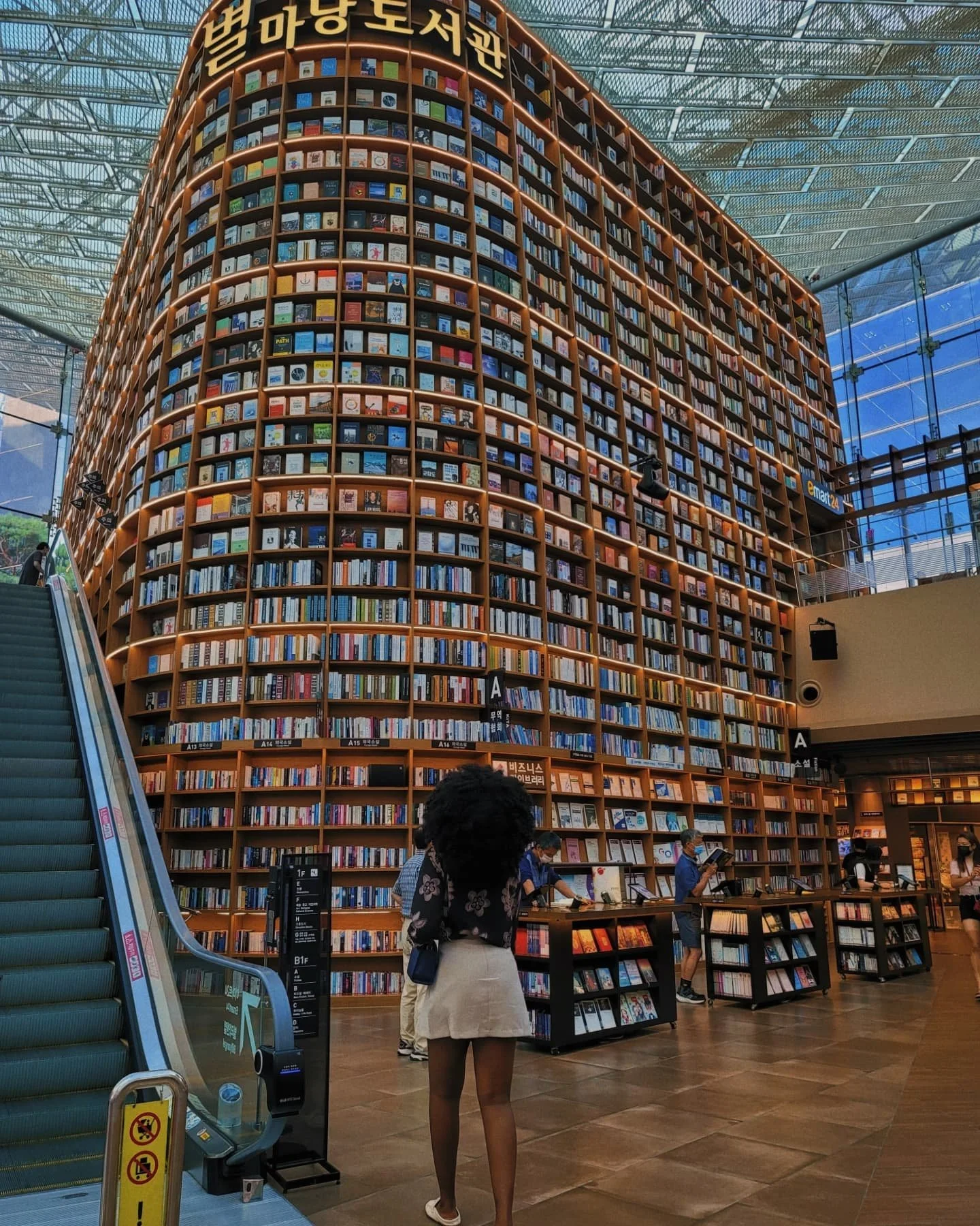 Interior of a modern bookstore with a large, curved wooden bookshelf filled with books. Several people browse and shop, with a woman in the foreground looking at books. An escalator is visible on the left side, and the ceiling is made of glass with metal framework.