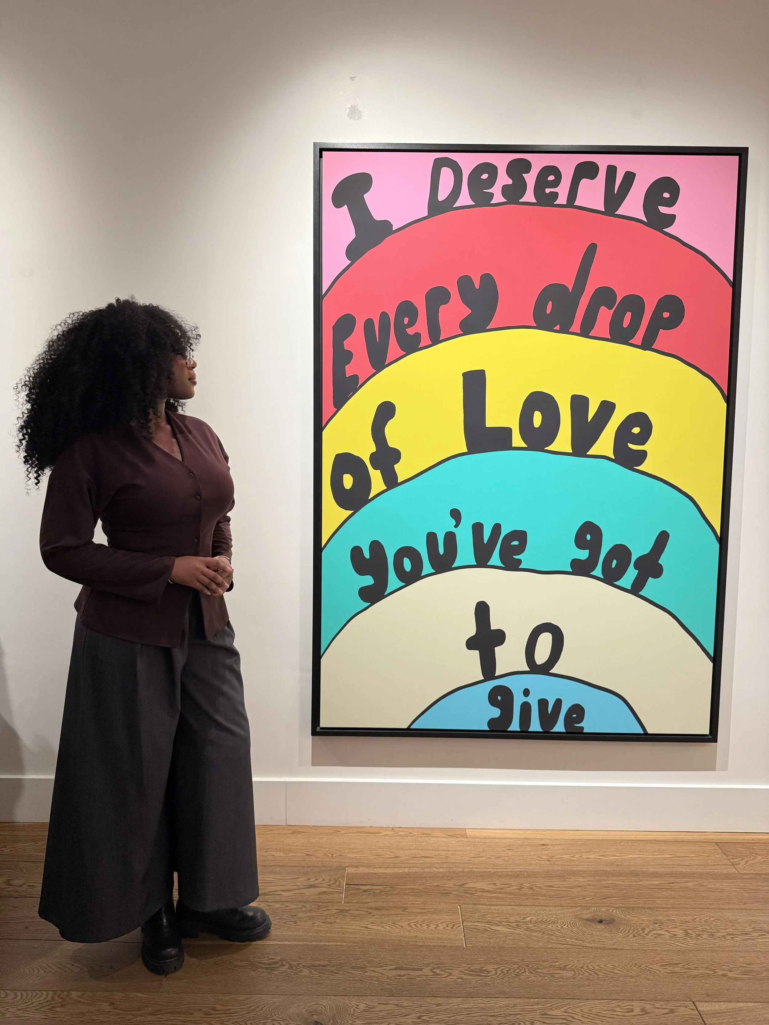 A woman with curly hair, wearing a dark brown blazer and dark wide-leg pants, stands in front of a large, colorful artwork with the words 'Deserve every drop of love you're' written in bold black letters on rainbow-colored arcs.