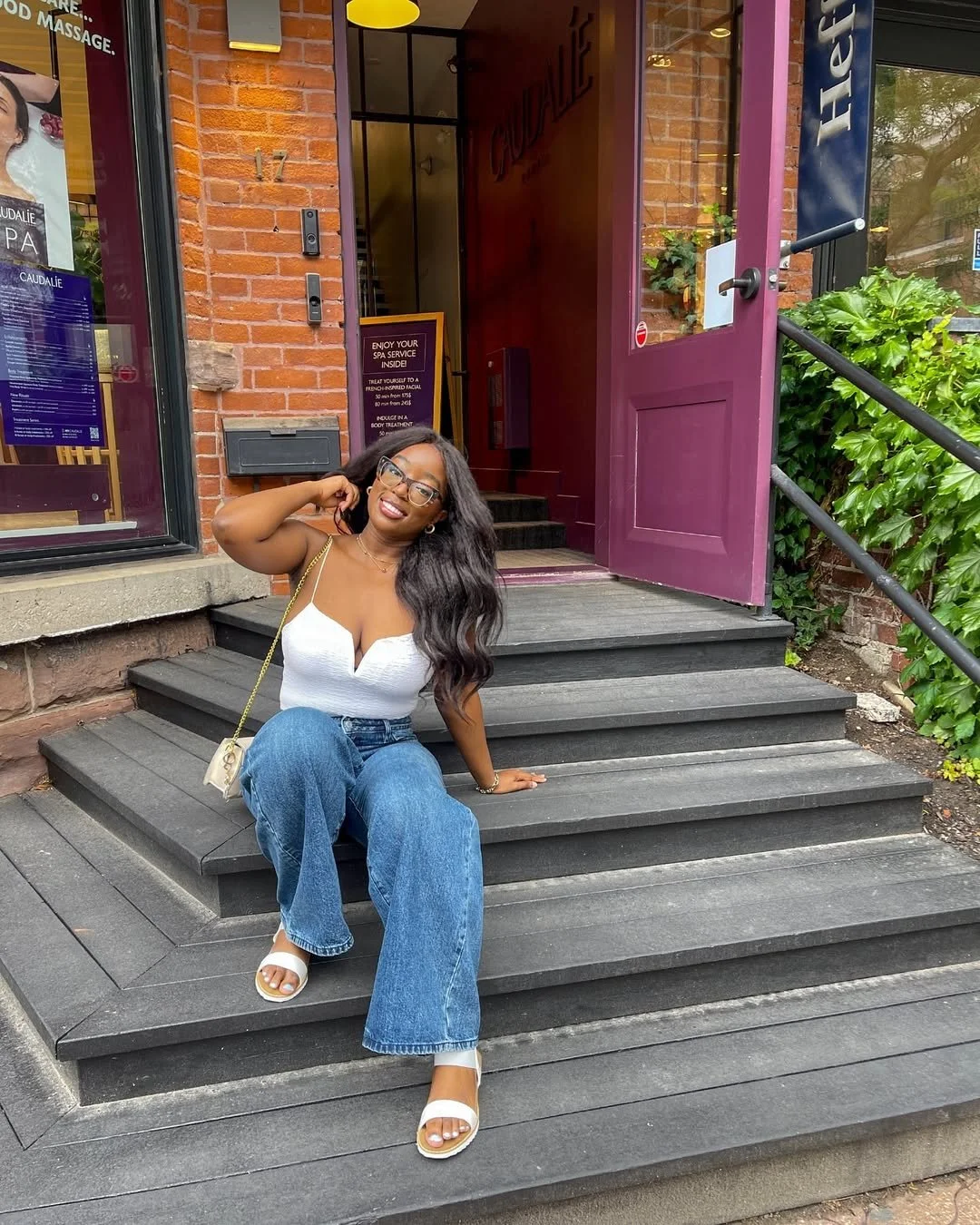 A woman with long dark hair, glasses, and a white tank top sitting on black wooden stairs outside a brick building with purple door, smiling and posing for the camera.
