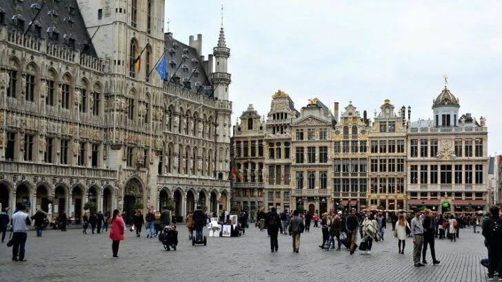 Crowd of people walking in a historic European square with ornate, old buildings and a castle-like structure on the left.