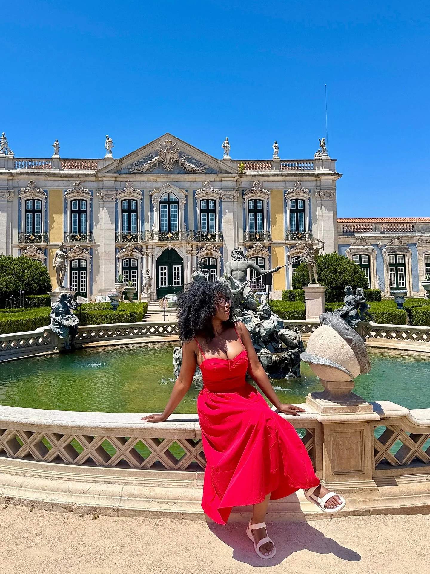 A woman in a red dress sitting on a stone ledge in front of a fountain with statues, in front of a large ornate building under a clear blue sky.
