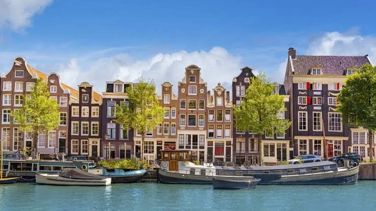 A row of traditional Dutch-style houses with gabled roofs along a canal, with boats docked in the water and green trees in front, under a partly cloudy blue sky.