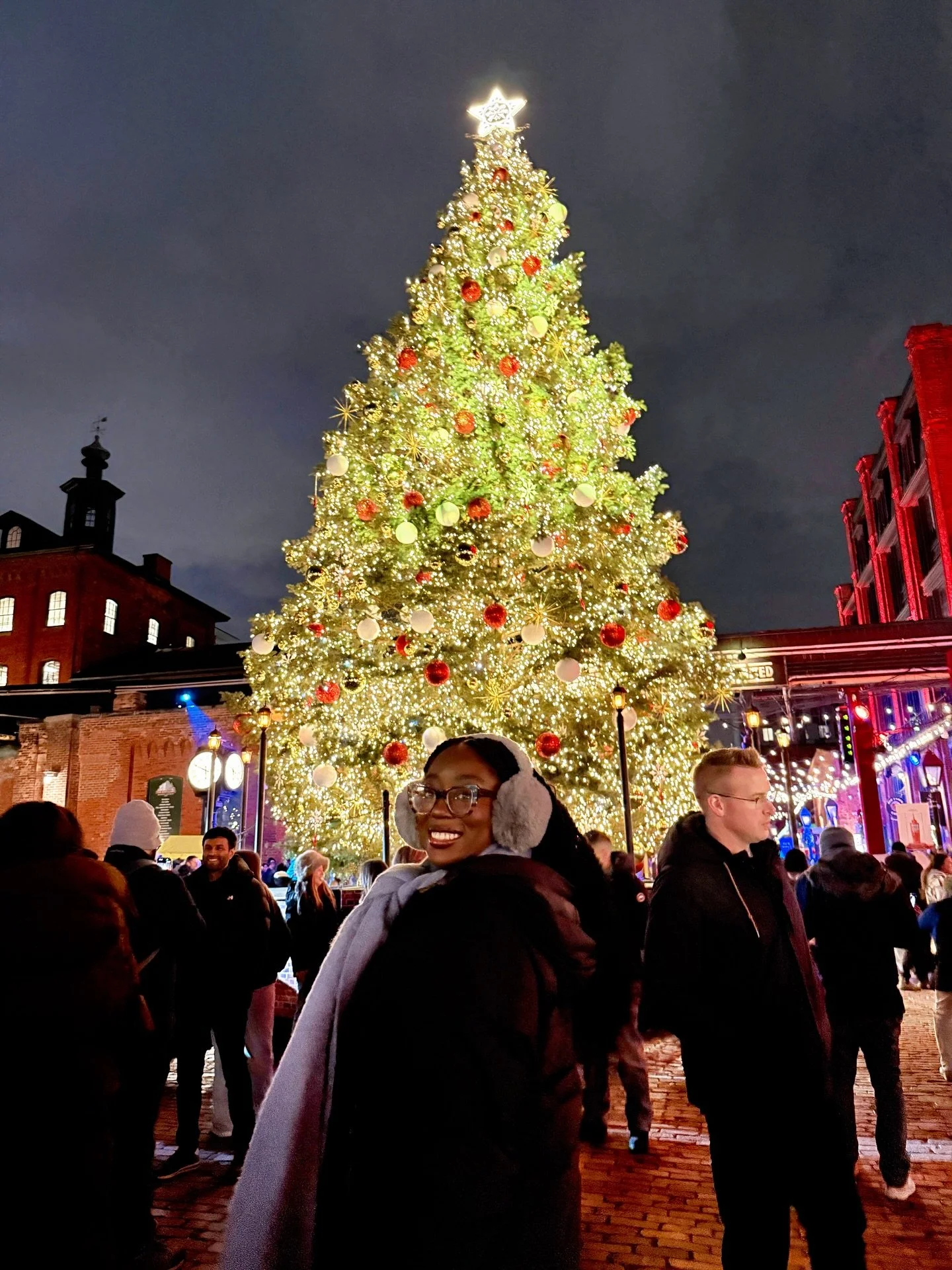 A large decorated Christmas tree with bright lights, red and white ornaments, and a star on top, in a crowded outdoor area at night with people smiling and standing in front of the tree.