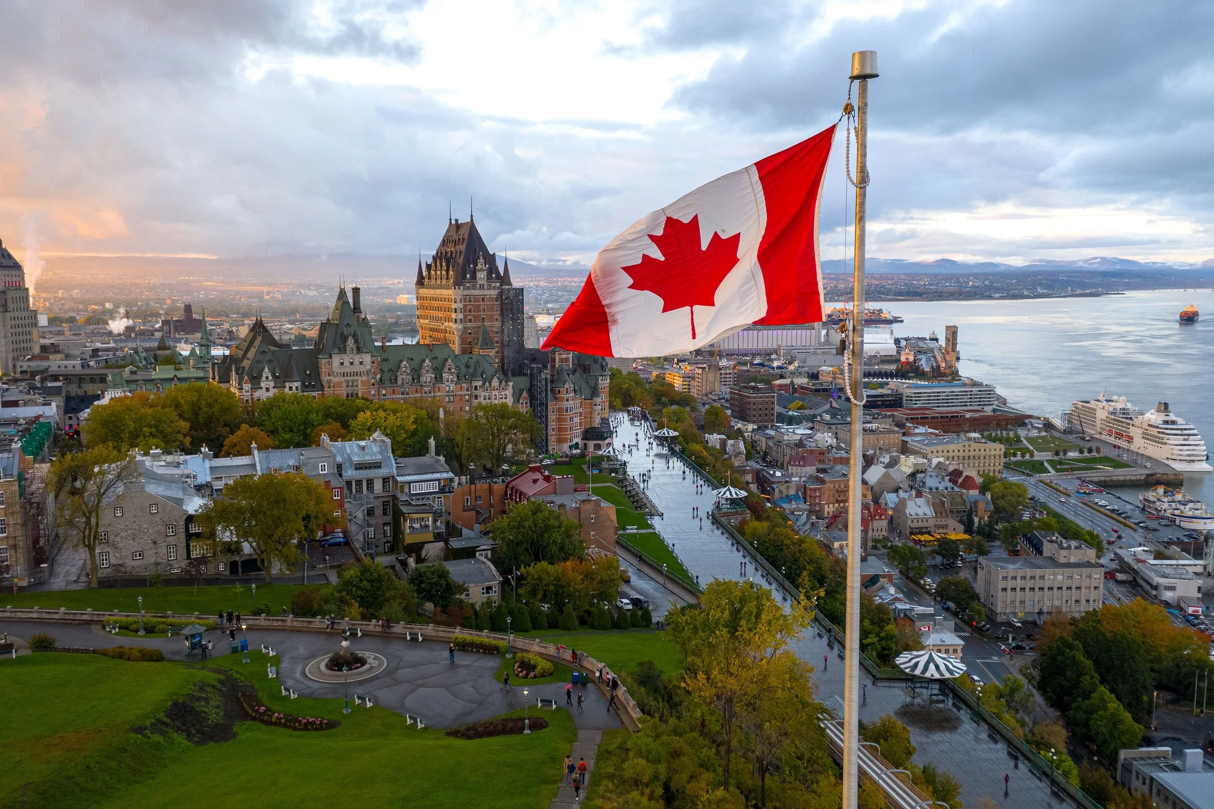 A city skyline with historic buildings, a river with boats, and a Canadian flag on a tall pole in the foreground. The sky is cloudy with patches of sunlight.