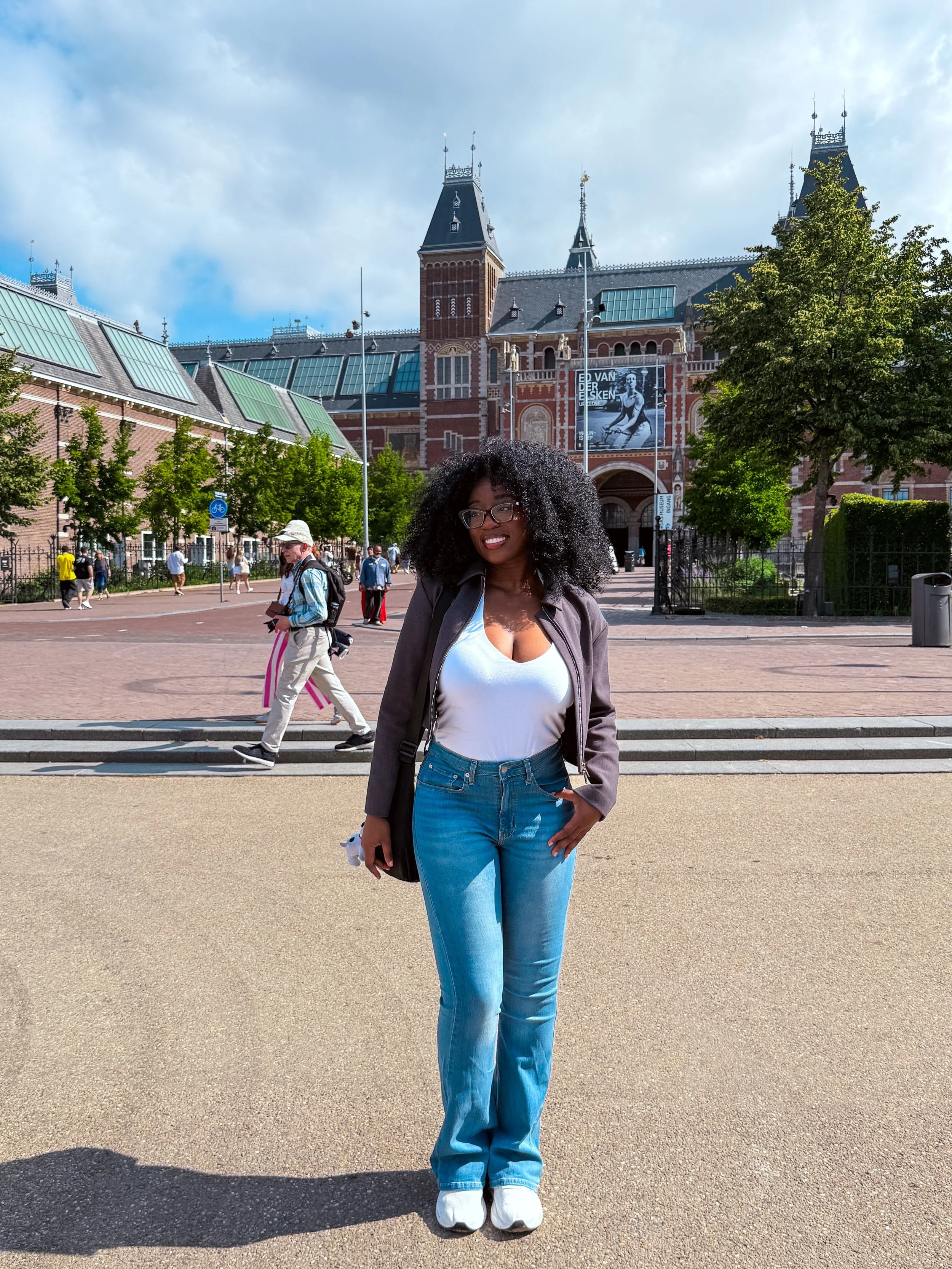 A woman with curly black hair, glasses, a white shirt, and blue jeans standing outside near the Rijksmuseum in Amsterdam, with the historic building and a tree in the background.