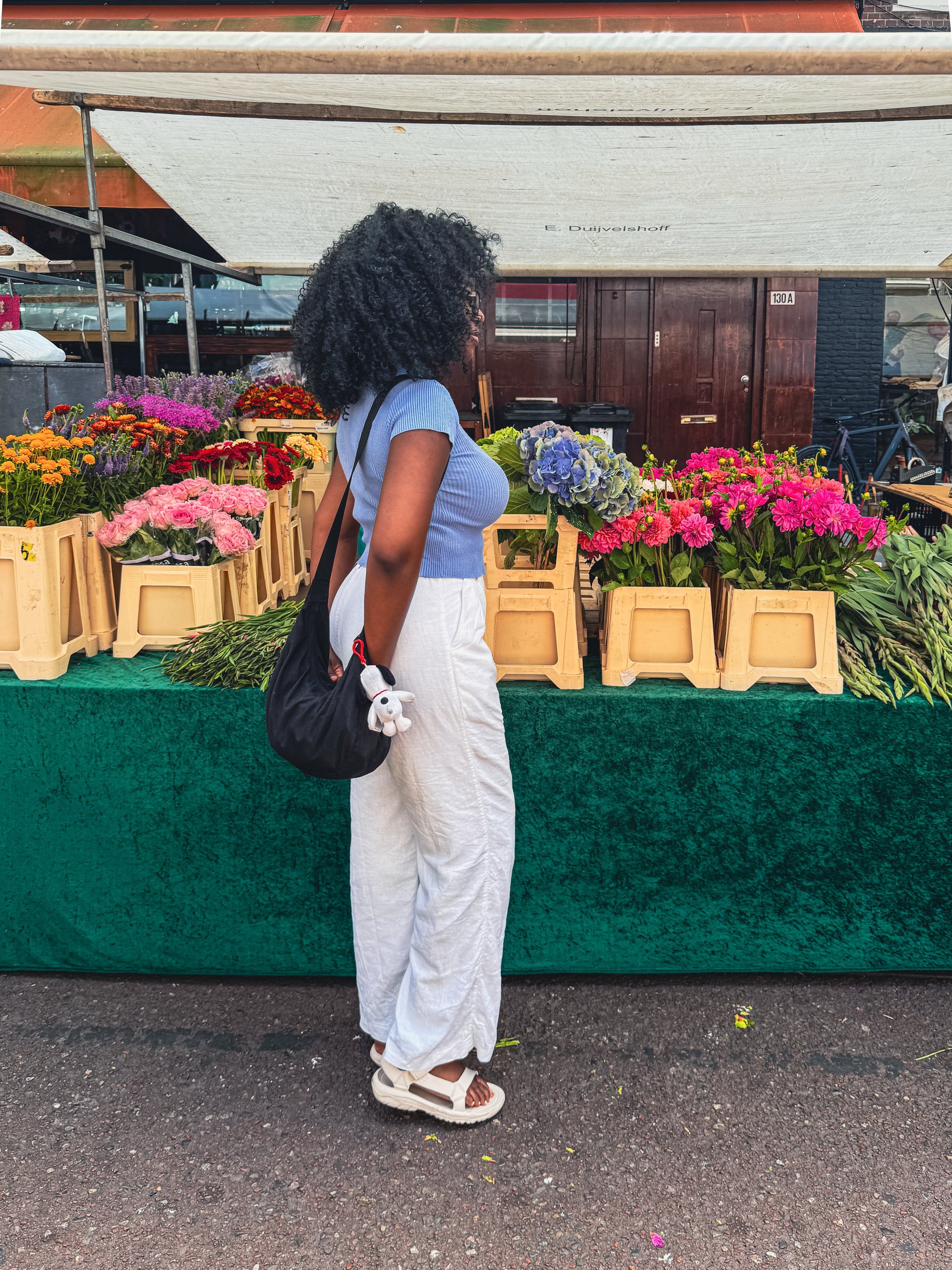 A woman standing in front of a flower stand at an outdoor market. She has curly black hair, glasses, and is wearing a light blue short-sleeve top, white wide-leg pants, and white sandals. She carries a black bag with a plush Snoopy keychain. The flower stand displays various colorful flowers in beige crates and has a green carpet covering the table. There is a dark brown building in the background.