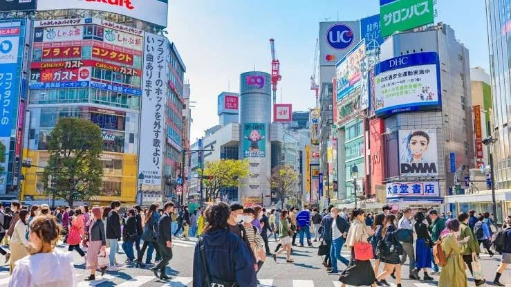 Crowded crosswalk in a busy city intersection with tall buildings covered in colorful billboards and signs, in what appears to be Tokyo, Japan.