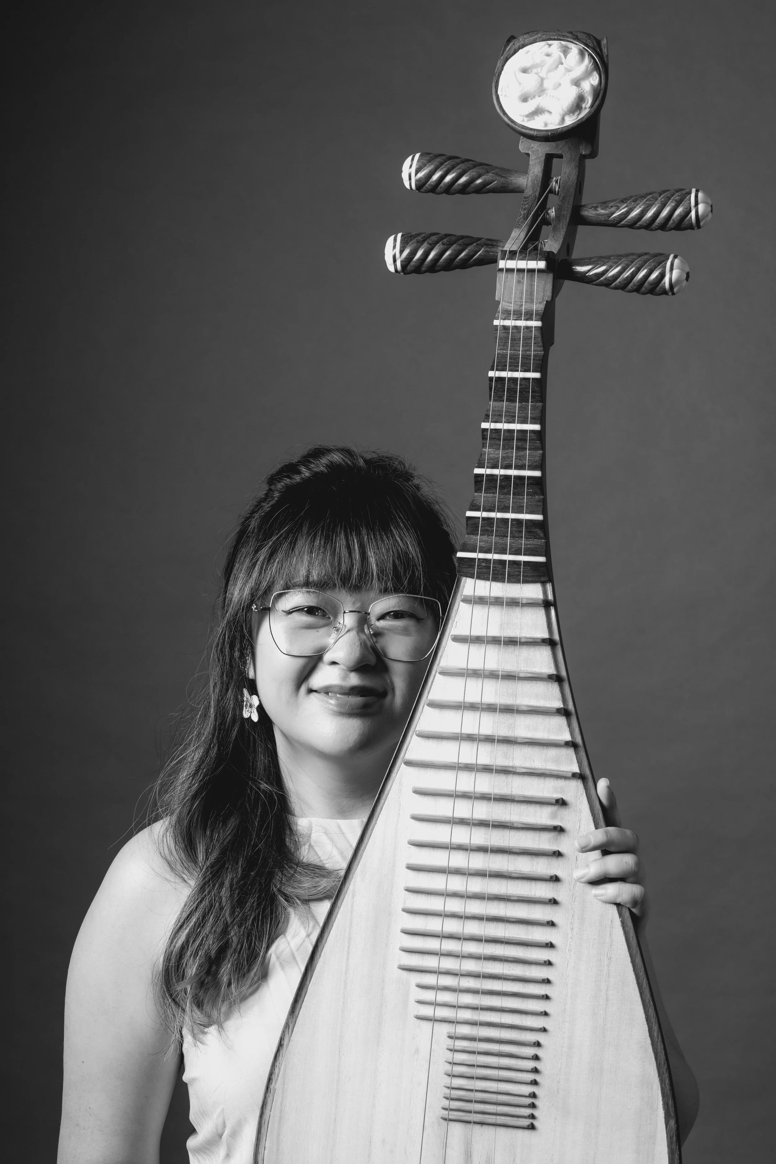 A woman with long hair and glasses smiling, holding a large, traditional stringed musical instrument, possibly a zither or similar, in a studio with a plain background.