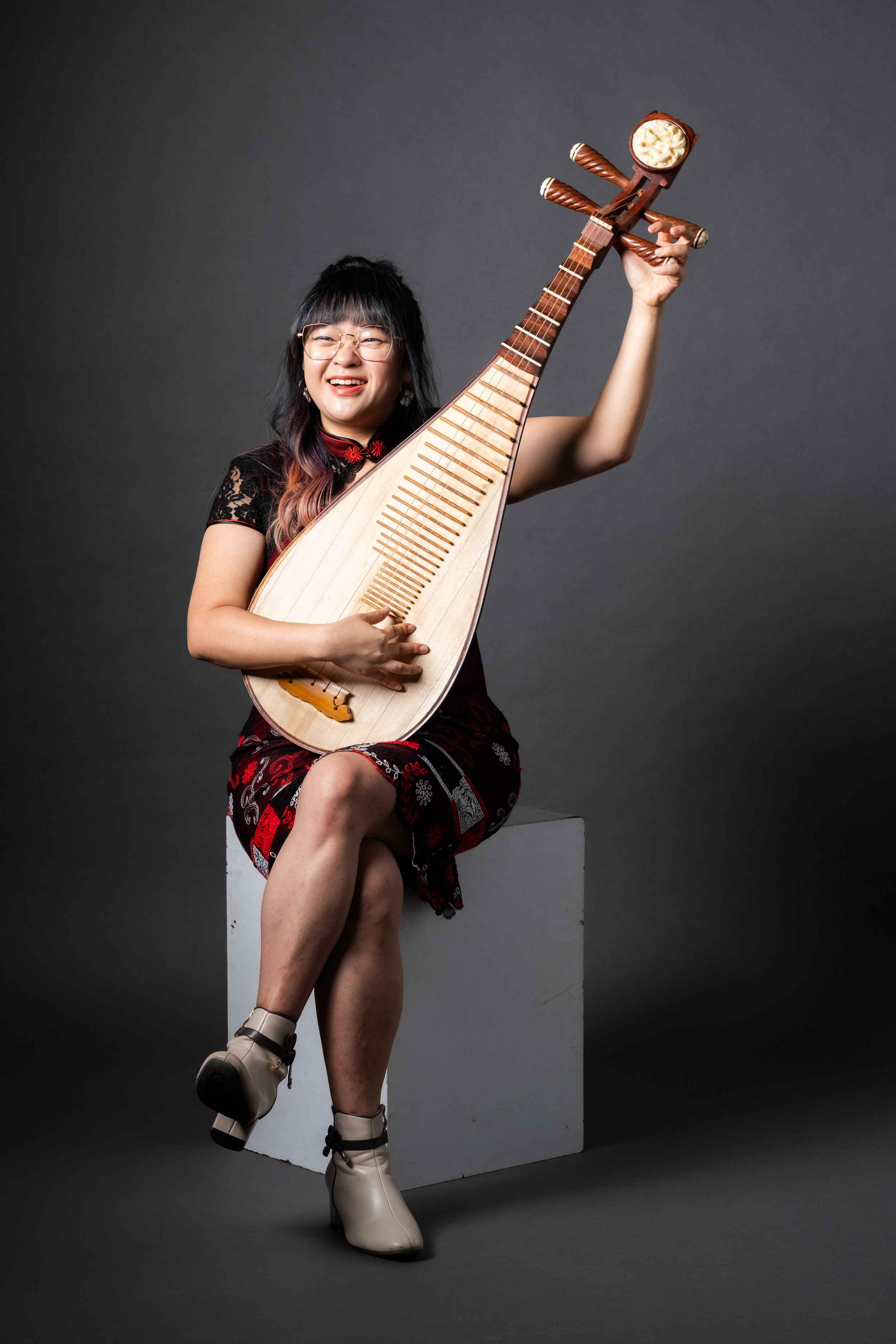A woman with glasses, dark hair with colored tips, wearing a black and red dress with floral patterns, sitting on a white block against a gray background. She is smiling and holding a traditional Asian musical instrument with a long fretboard and a head resembling a bowl of whipped cream, raising it in her right hand. Her left leg is crossed over her right, and she wearing beige high-heeled boots with laces.