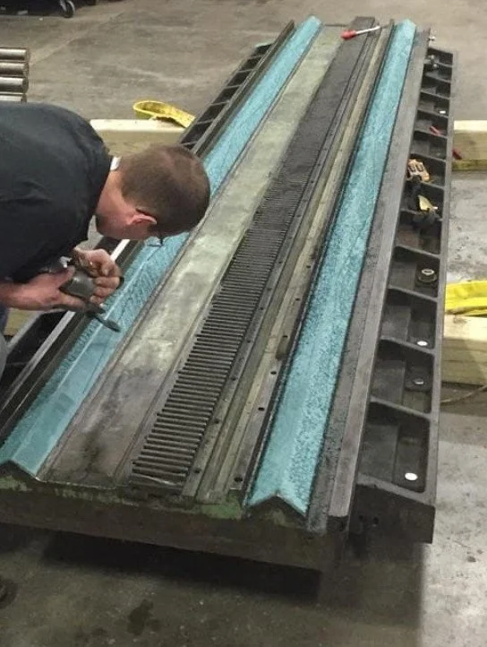 A man working on a train track model or a track fixture, using a power tool. The setup is on a large black workbench in an industrial workshop setting.