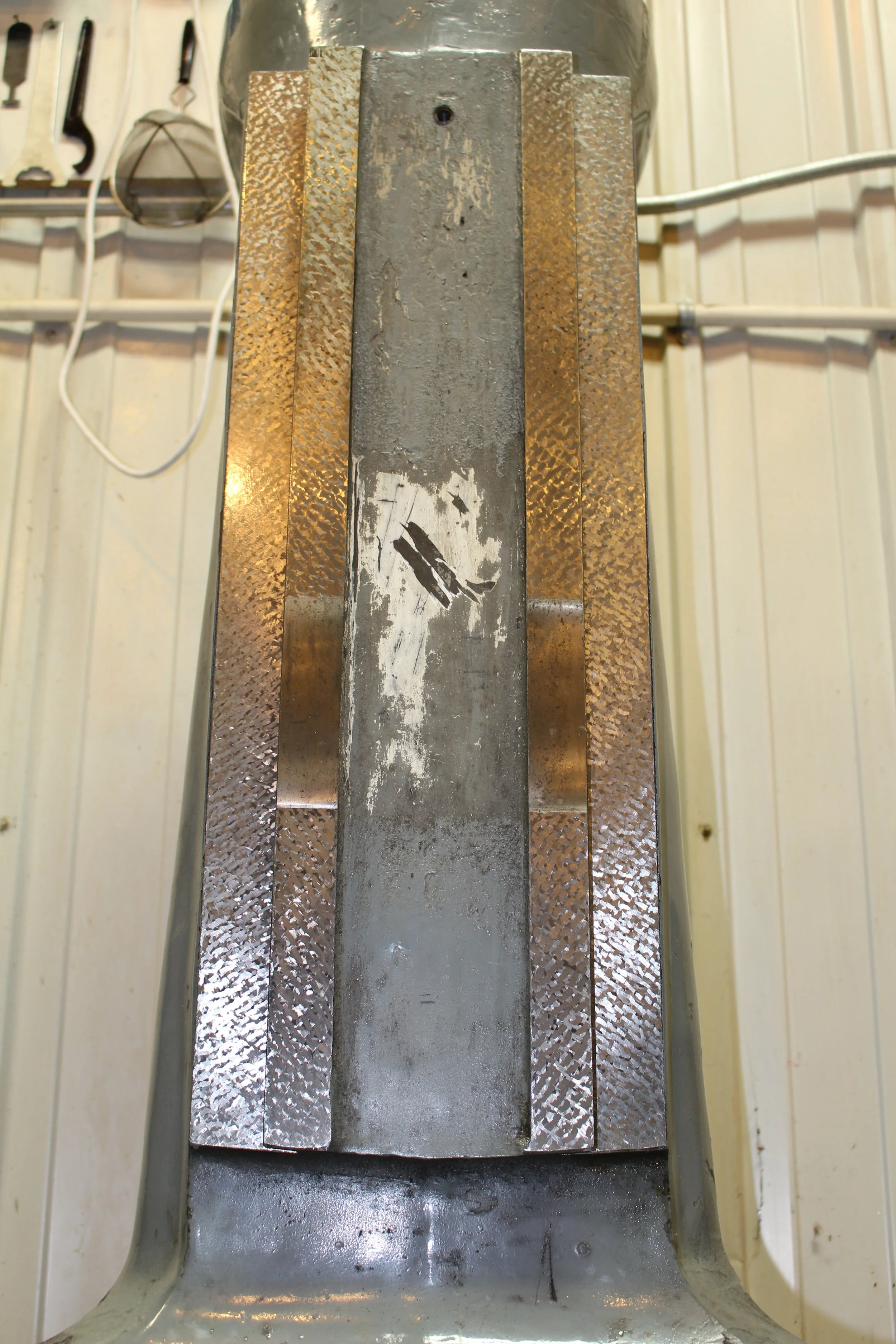 Close-up of a metal industrial or scientific vent or duct with a cylindrical shape and a silver color, mounted inside a beige or cream-colored wall with vertical paneling. The interior surfaces of the vent show signs of wear and scratches.