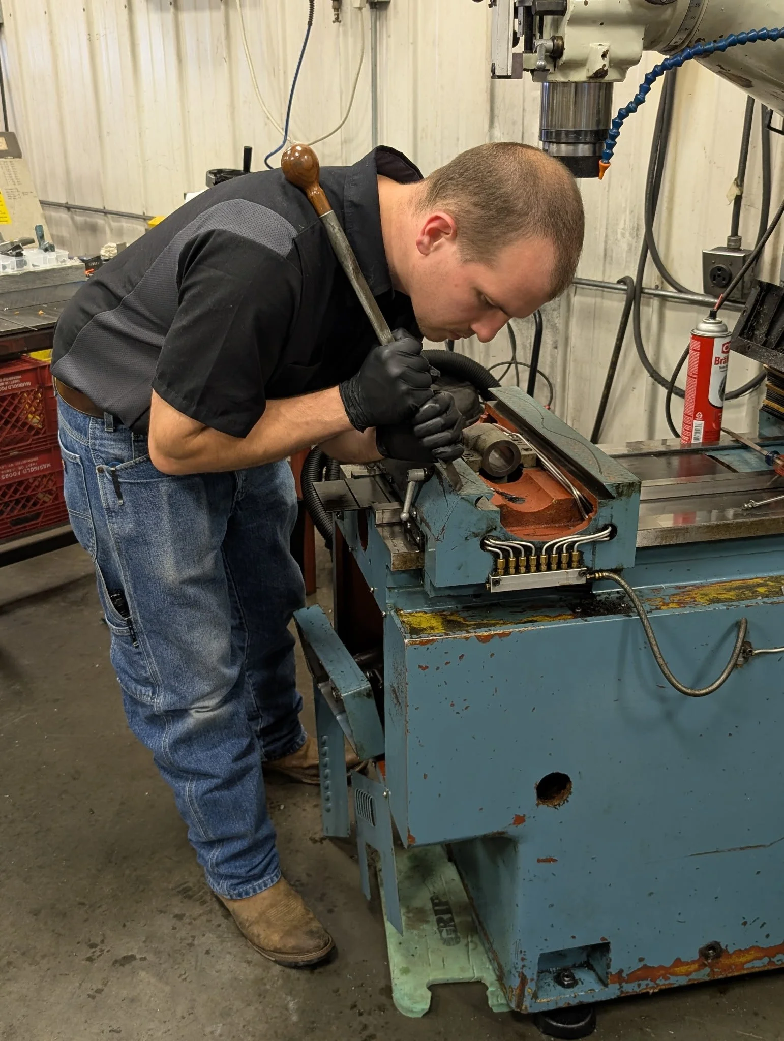 Man wearing black gloves and jeans working on a large blue industrial machine in a workshop, with tools and equipment around him.