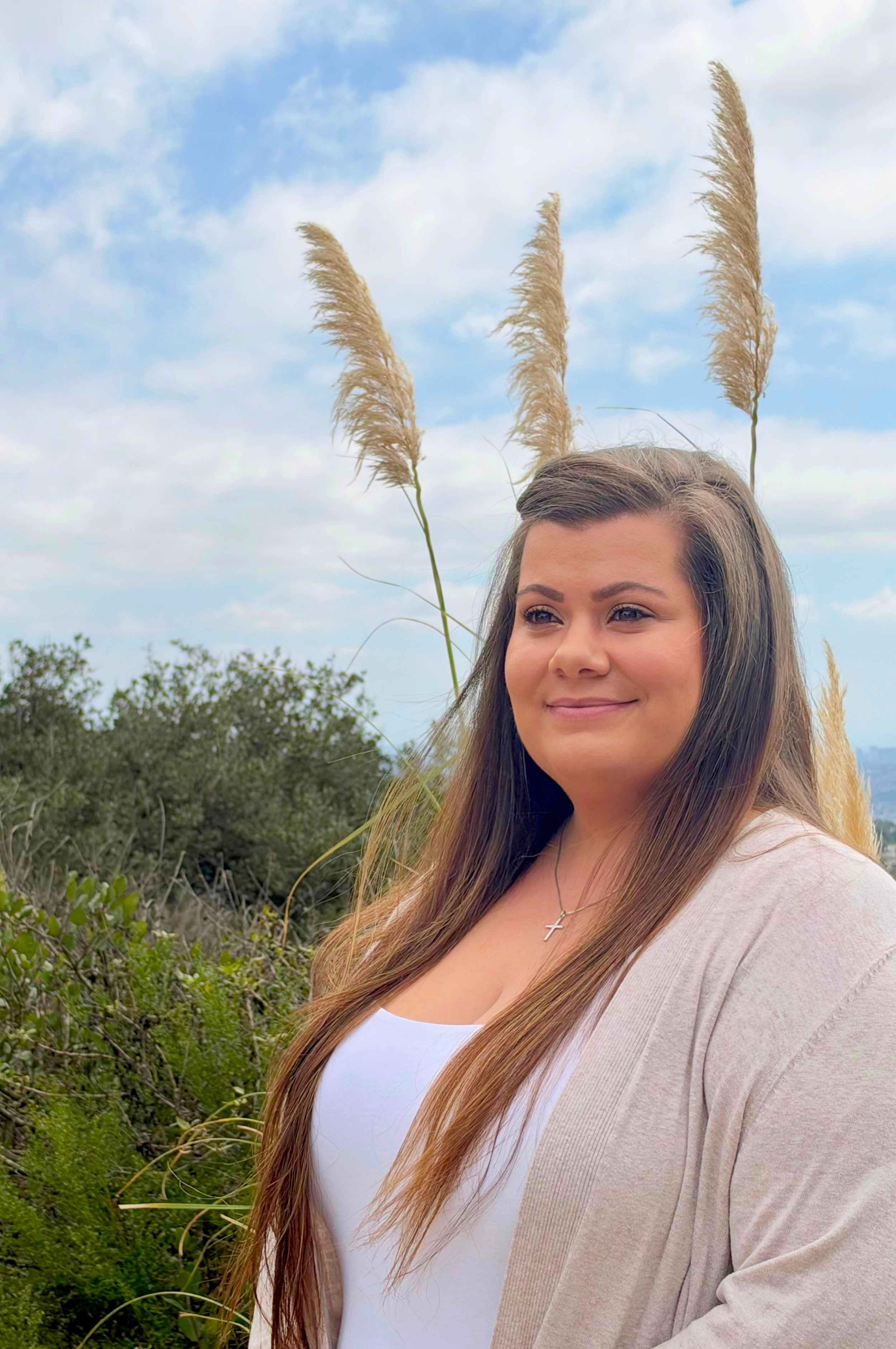 Founder - A woman with long brown hair wearing a white shirt and beige cardigan standing outdoors in a natural setting with green bushes and plants, with a cloudy sky in the background.